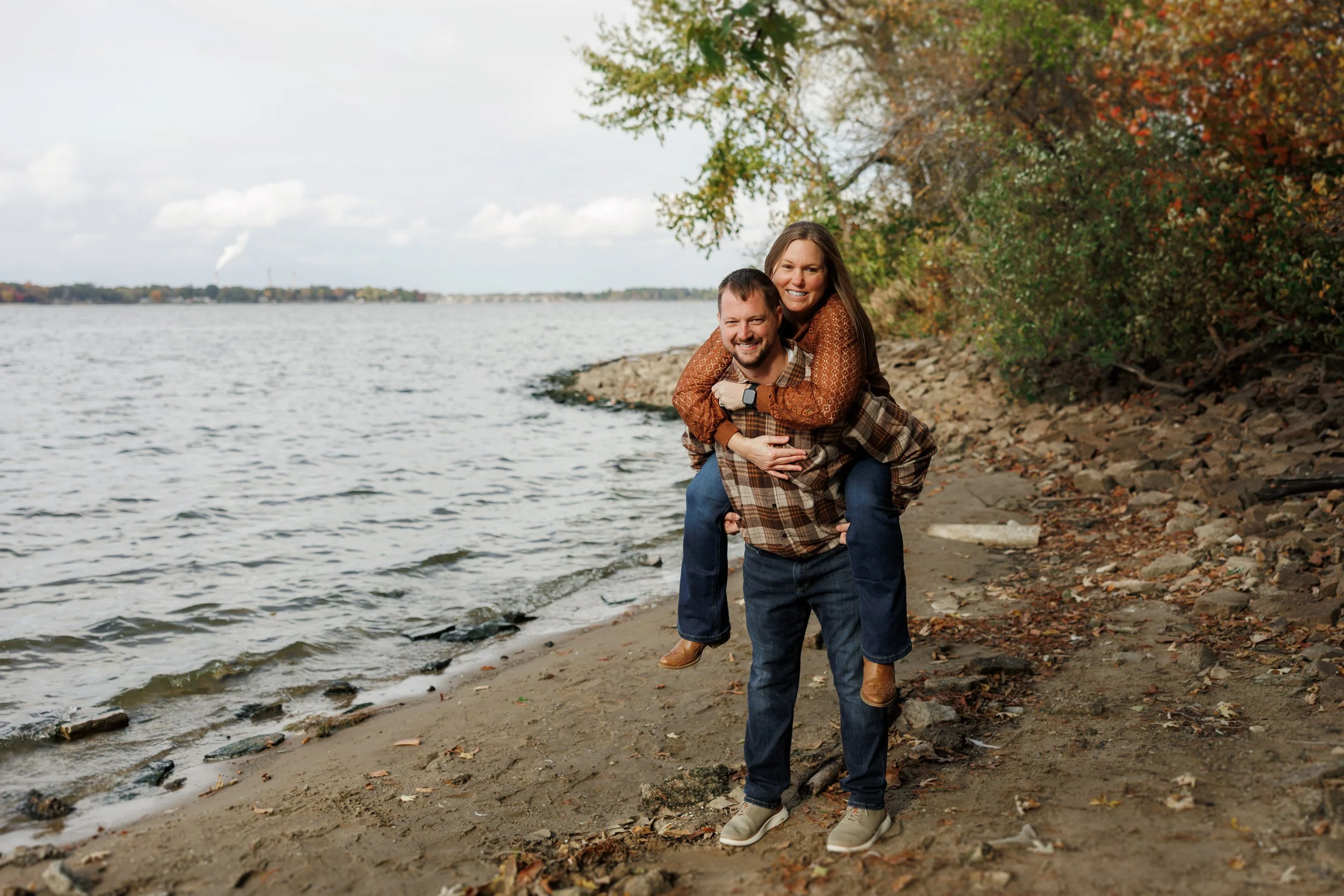 A man giving a woman a piggyback ride on a sandy beach with rocks and trees, with water and a cloudy sky in the background.