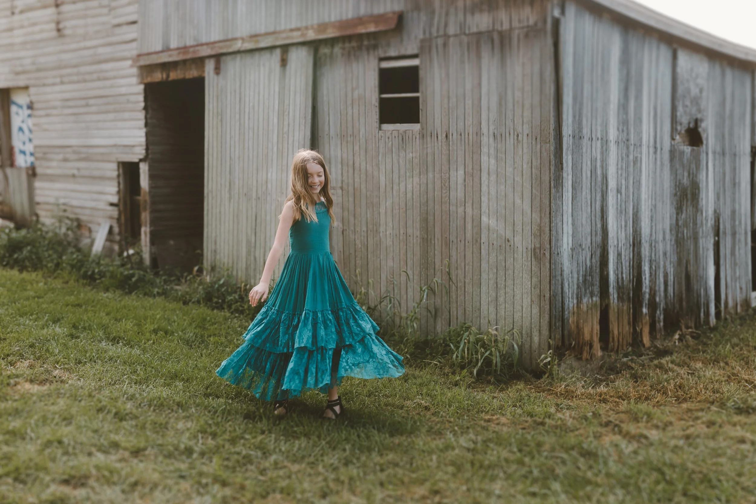 A young girl in a long blue dress walking on green grass next to a weathered wooden barn.