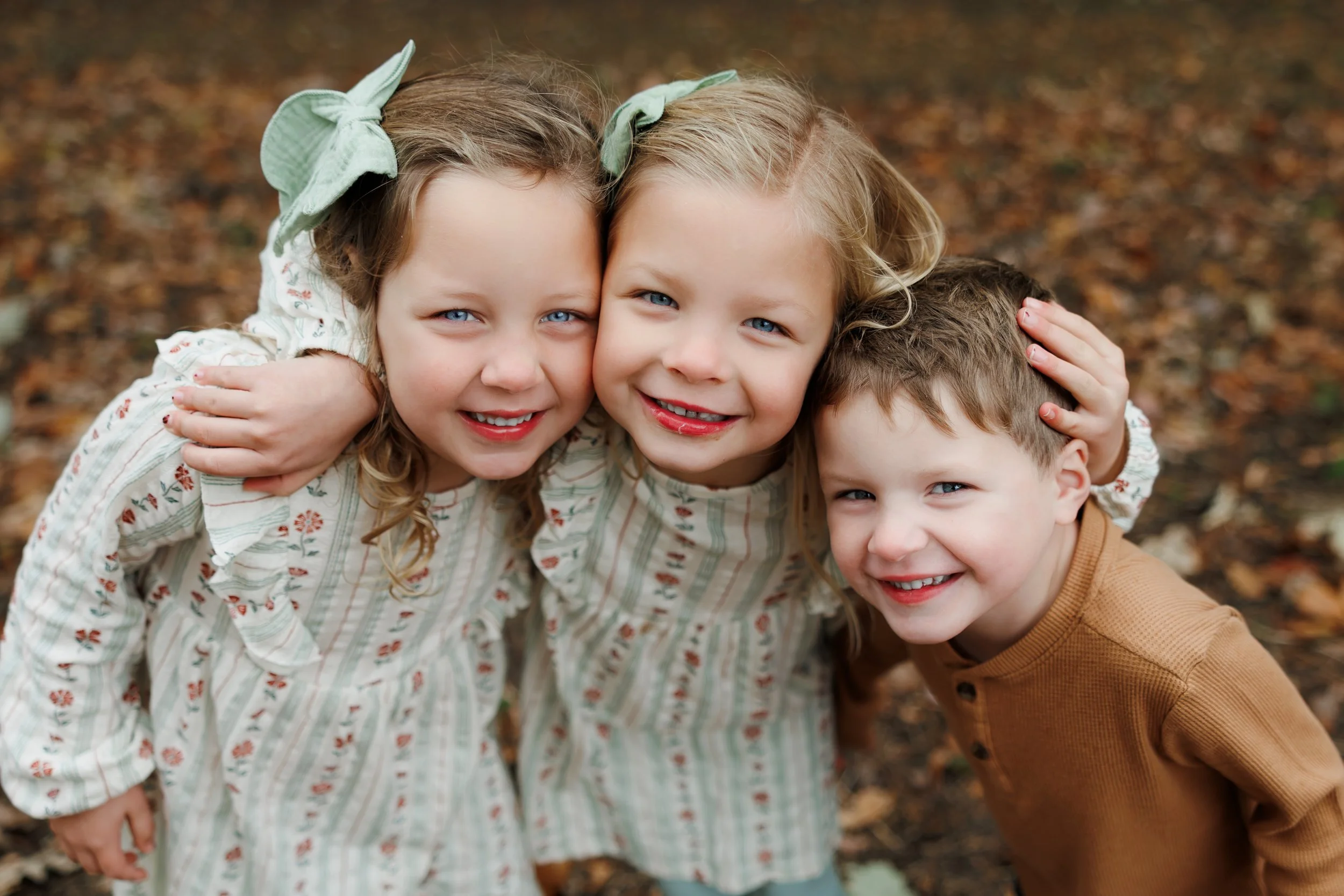 Three children hugging and smiling outdoors on a fall day with leaves on the ground.