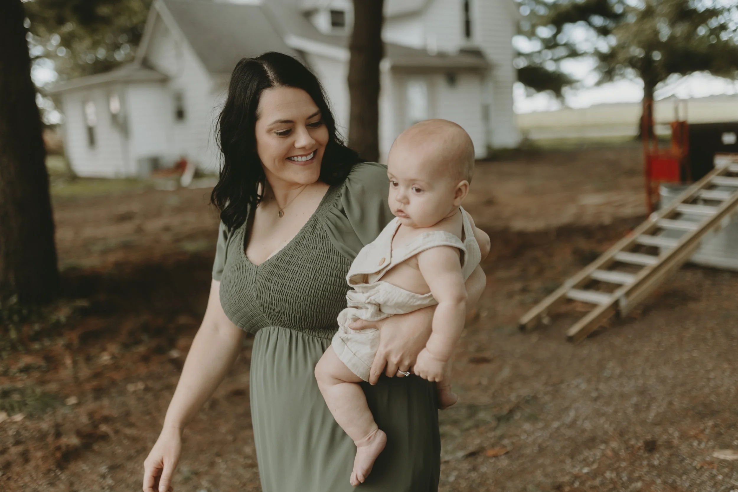 A woman with dark hair wearing a green dress is holding a baby outside near a house with trees in the background.