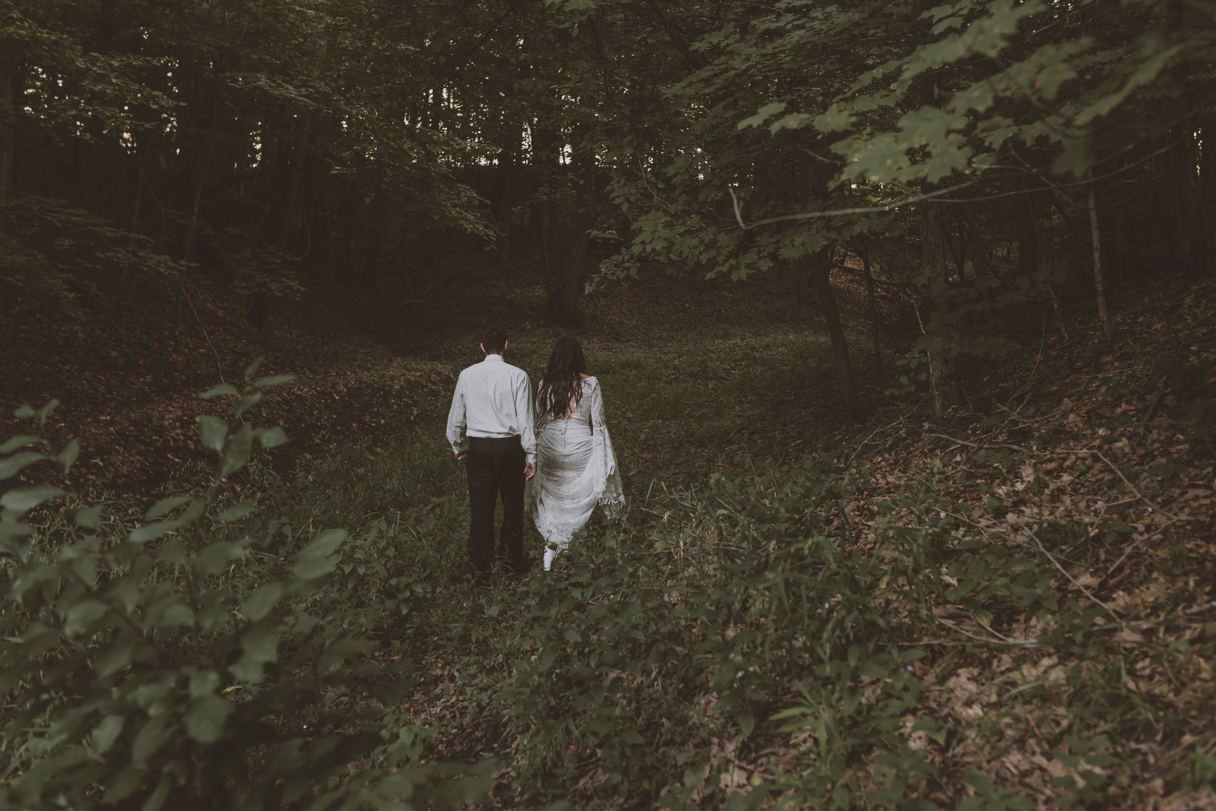 A couple walking hand-in-hand through a dense, dark forest during daytime, with trees and leaves surrounding them.