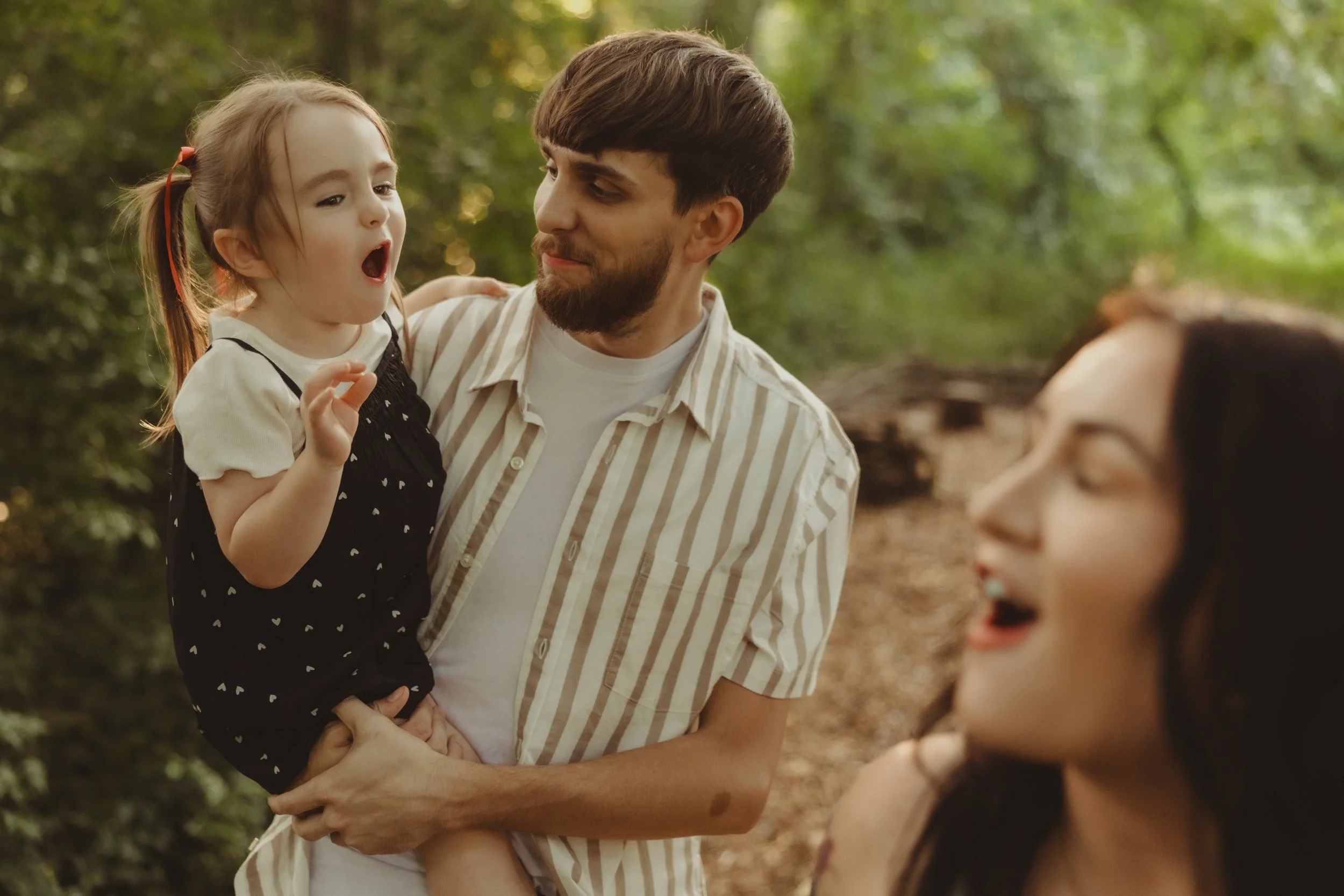 A man holding a young girl in his arms, both outdoors in a wooded area, with another girl partially visible in the foreground.
