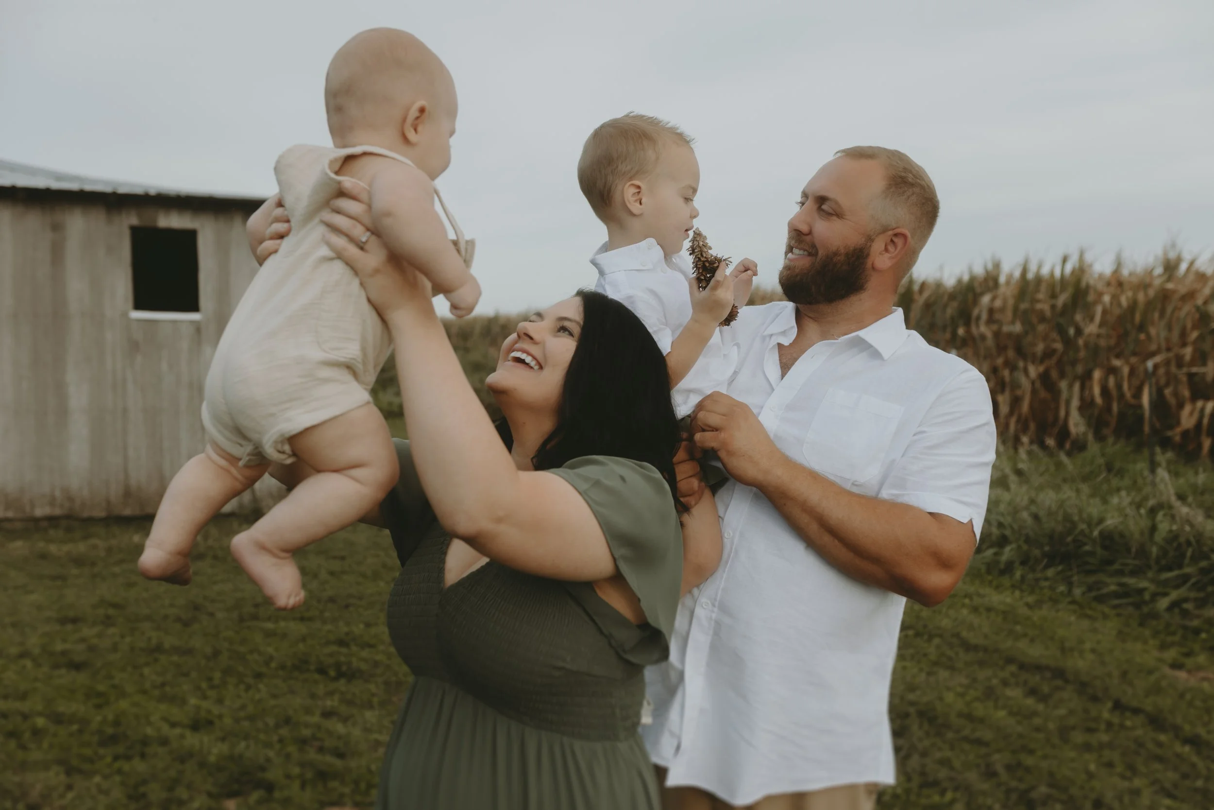 A family of four outdoors, the mother lifting a baby, and the father holding a young boy. The mother is smiling at the father, who is smiling back. The little boy is holding a pine cone and looking at the father. The background shows a barn and a field.