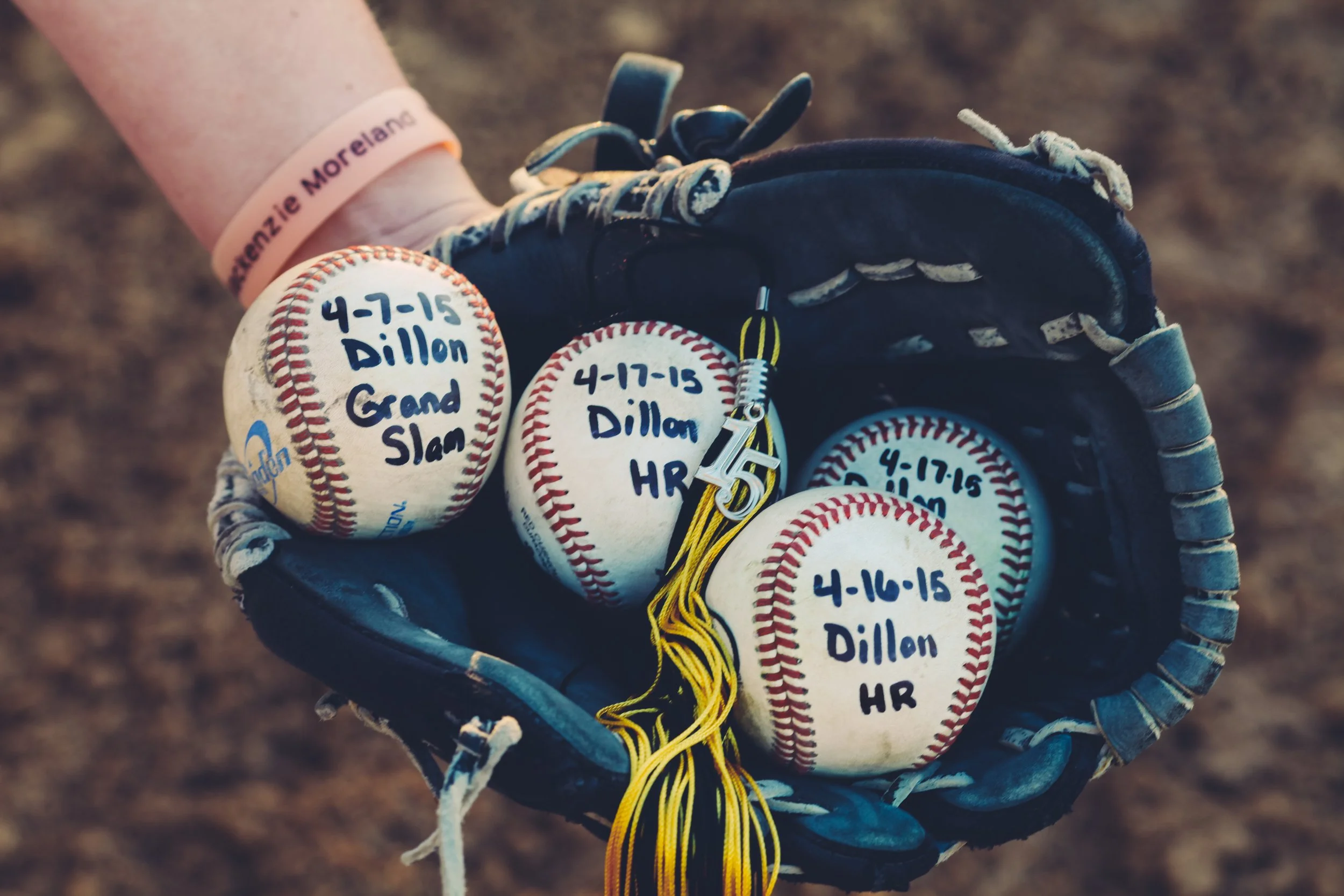 A baseball glove holding several baseballs with handwritten inscriptions showing dates and a nickname, on a dirt field.
