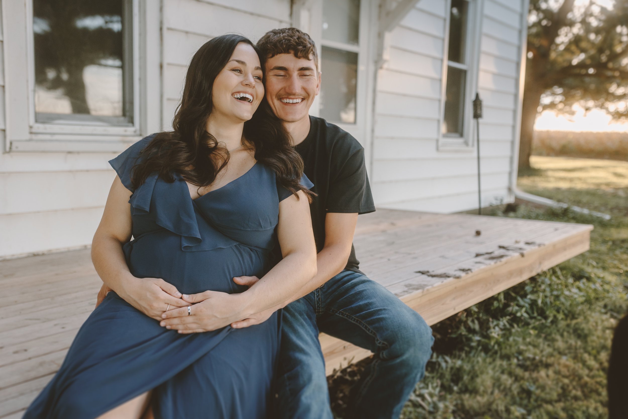 A smiling pregnant woman in a blue dress sitting on a man's lap on a porch, both looking happy and laughing.