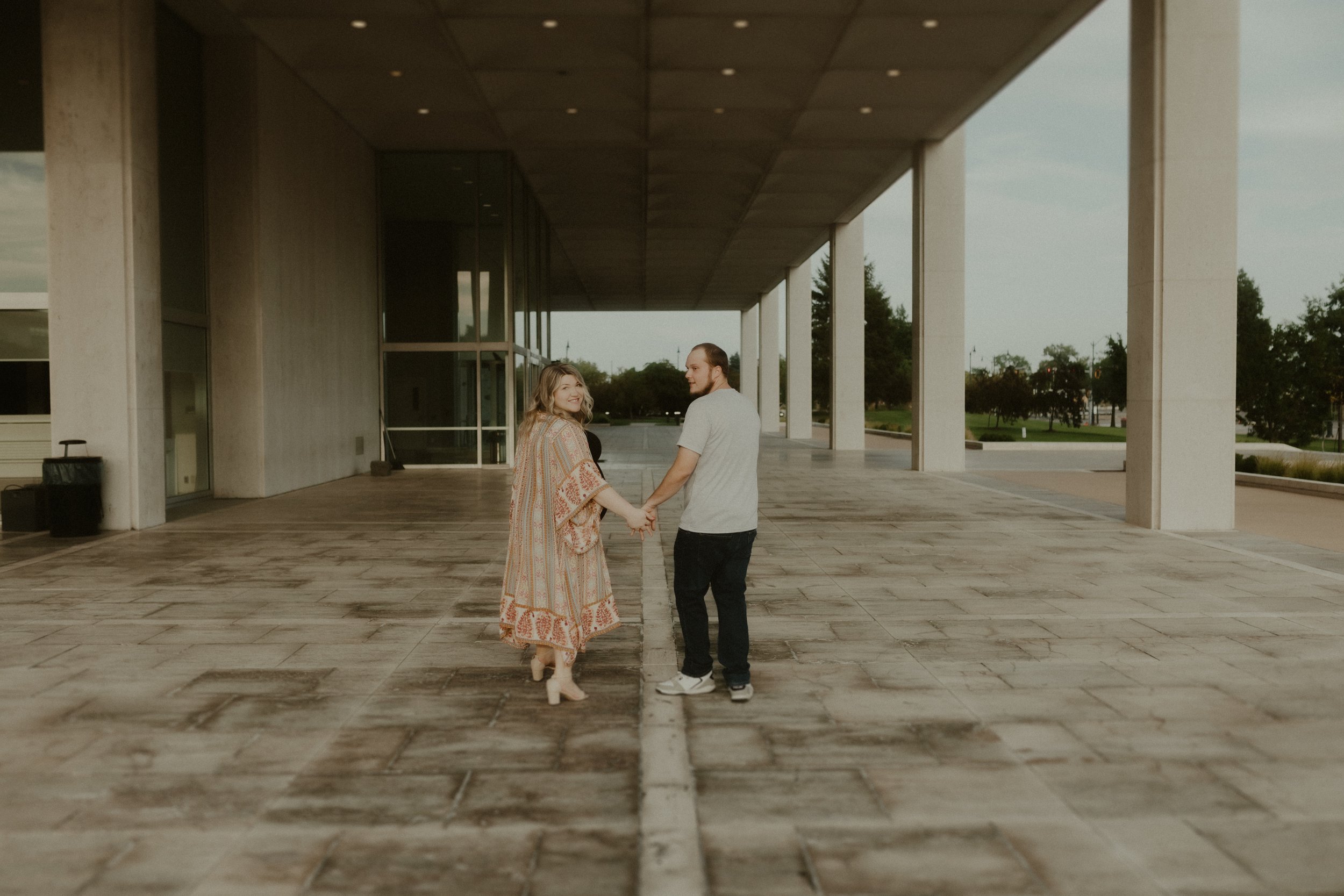 A couple holding hands and smiling, walking under a modern building with large columns and open space, with trees in the background.
