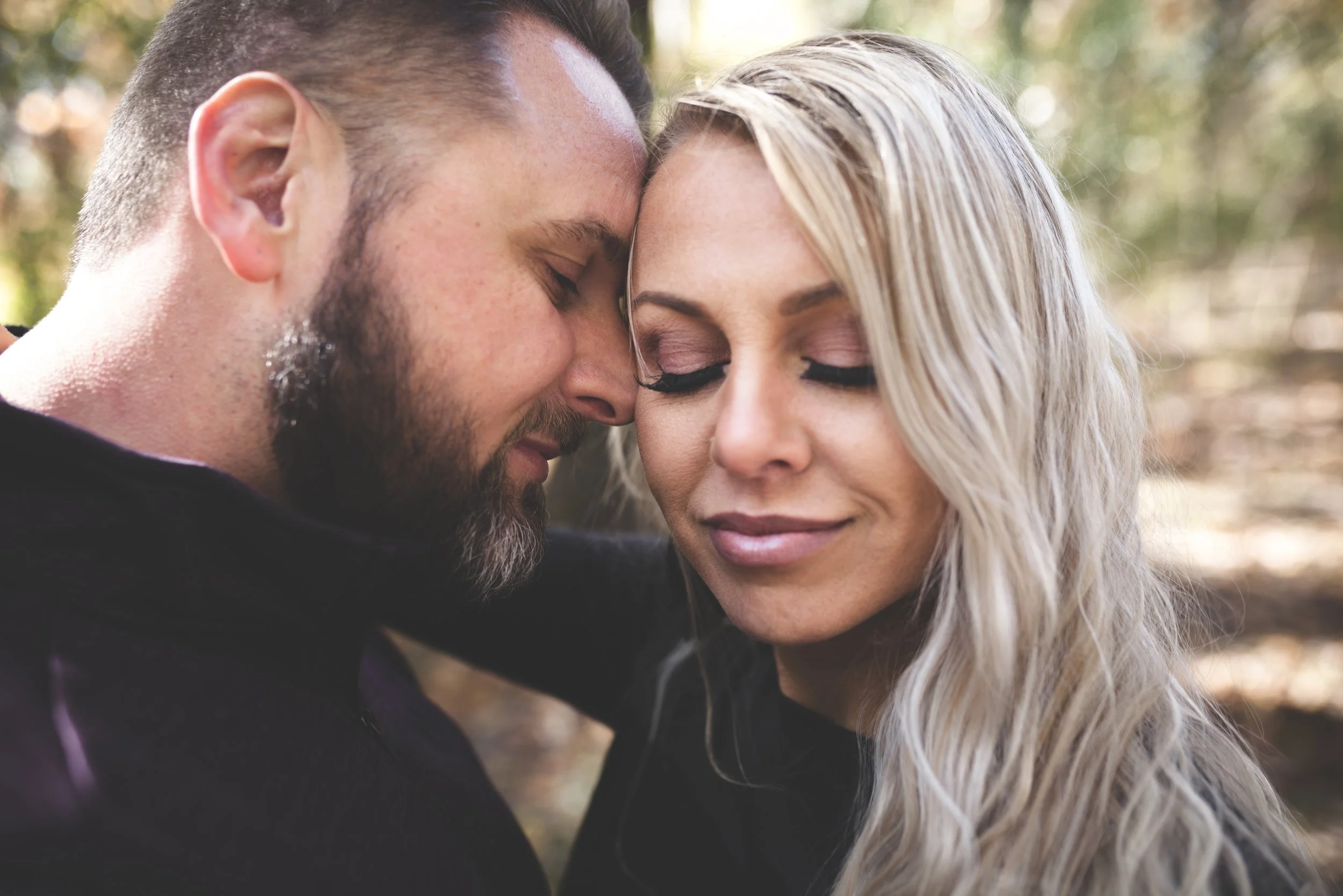 A close-up of a man and woman with their foreheads touching, eyes closed, sharing an intimate moment outdoors with blurred trees in the background.