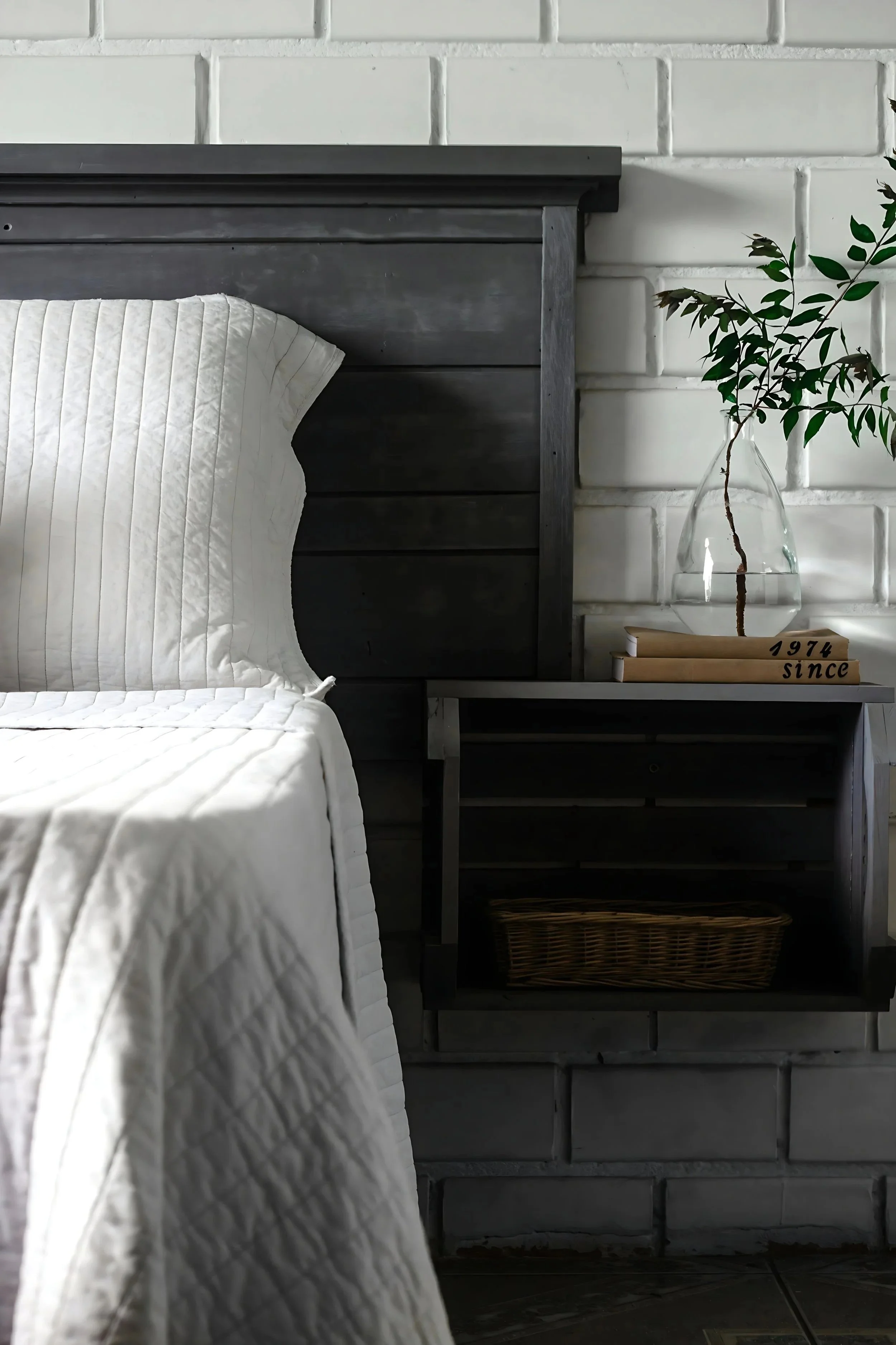 A bedroom with a dark gray wooden headboard, a white quilted bedspread, and a bedside table holding a glass vase with a branch and leaves, along with two books, one of which has '1974 since' written on it.