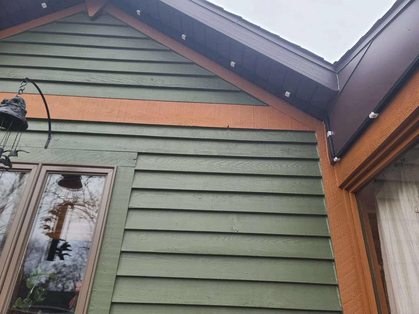 Close-up view of house exterior showing green horizontal siding, orange trim, a window with curtains, and part of the roof with solar panels. Task-Smart outdoor light install in Poinciana Florida.