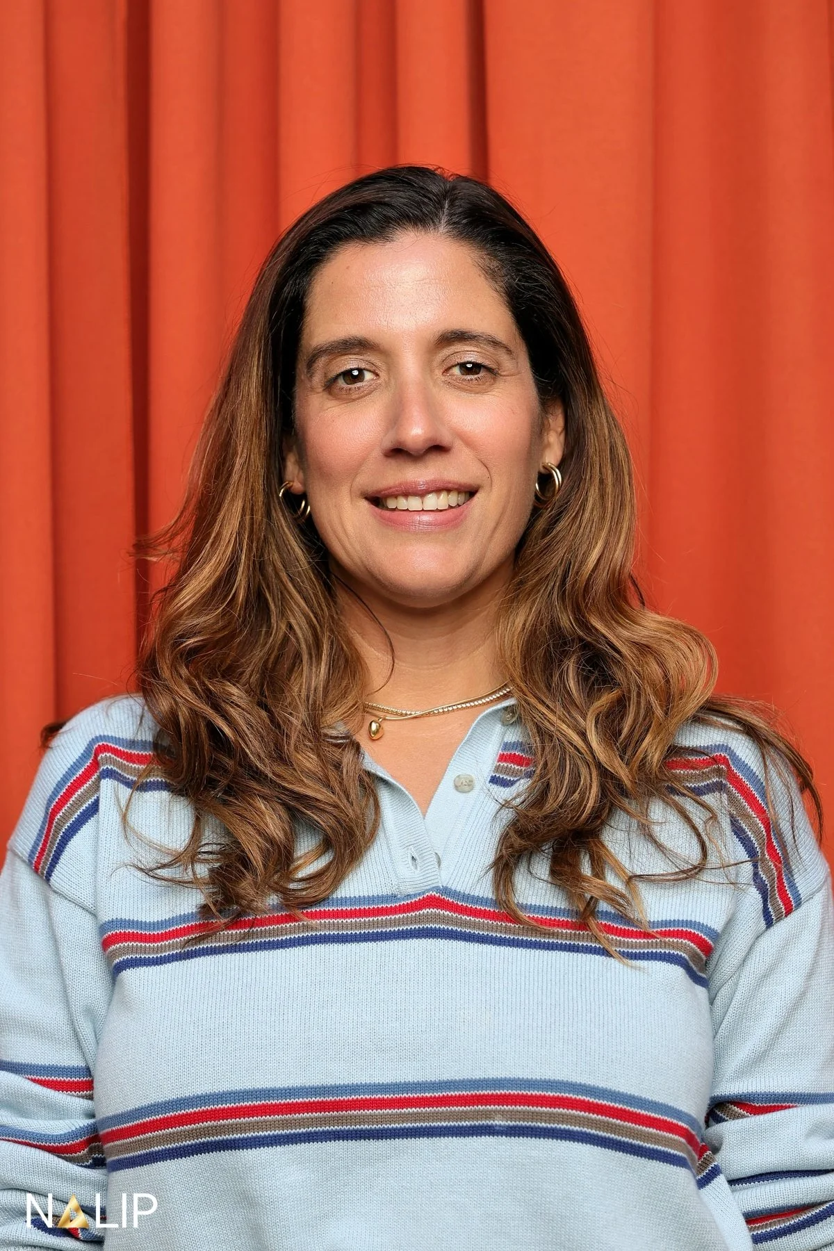 A woman with long wavy brown hair wearing a light blue striped sweater and gold jewelry standing in front of a red curtain.