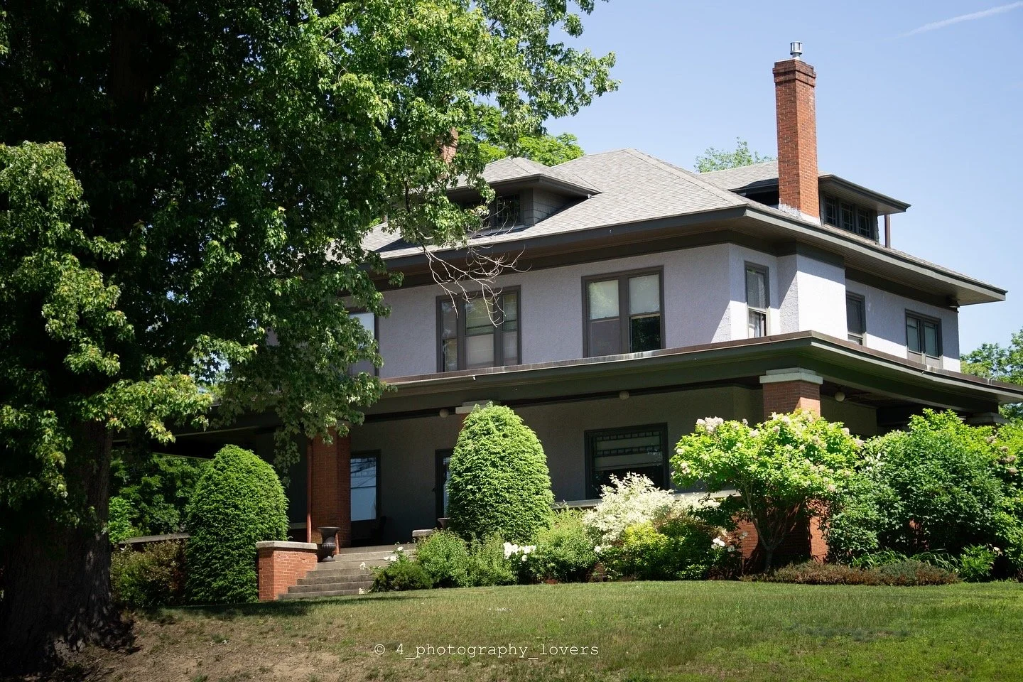 A large gray house with a front porch, surrounded by green trees and shrubs, under a clear blue sky.