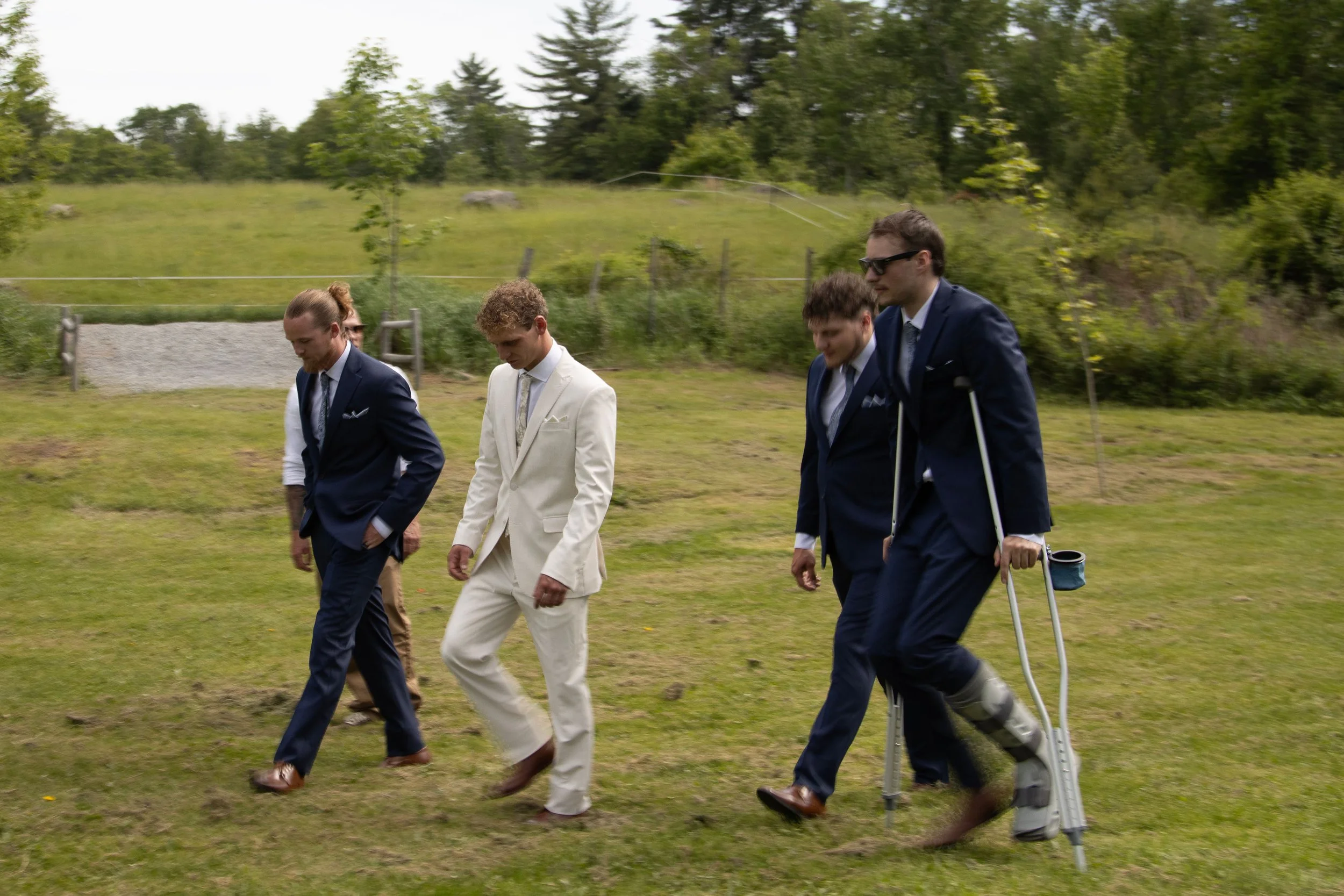 Four men in suits walking outdoors on grass, one with crutches, amidst green trees and a fence.