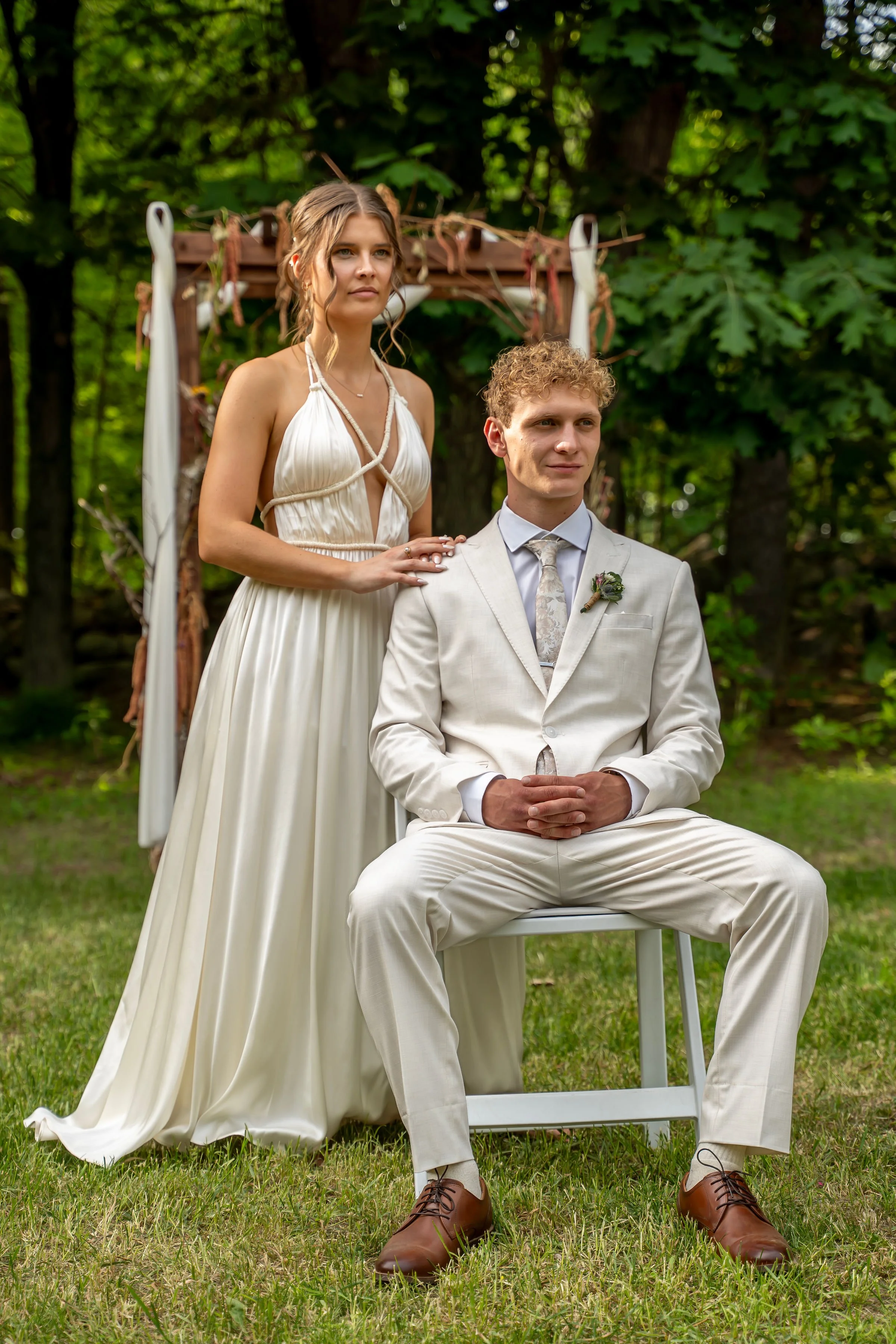 A young woman in a white dress and a young man in a white suit during an outdoor wedding ceremony in a forest setting, with a decorative arch in the background.