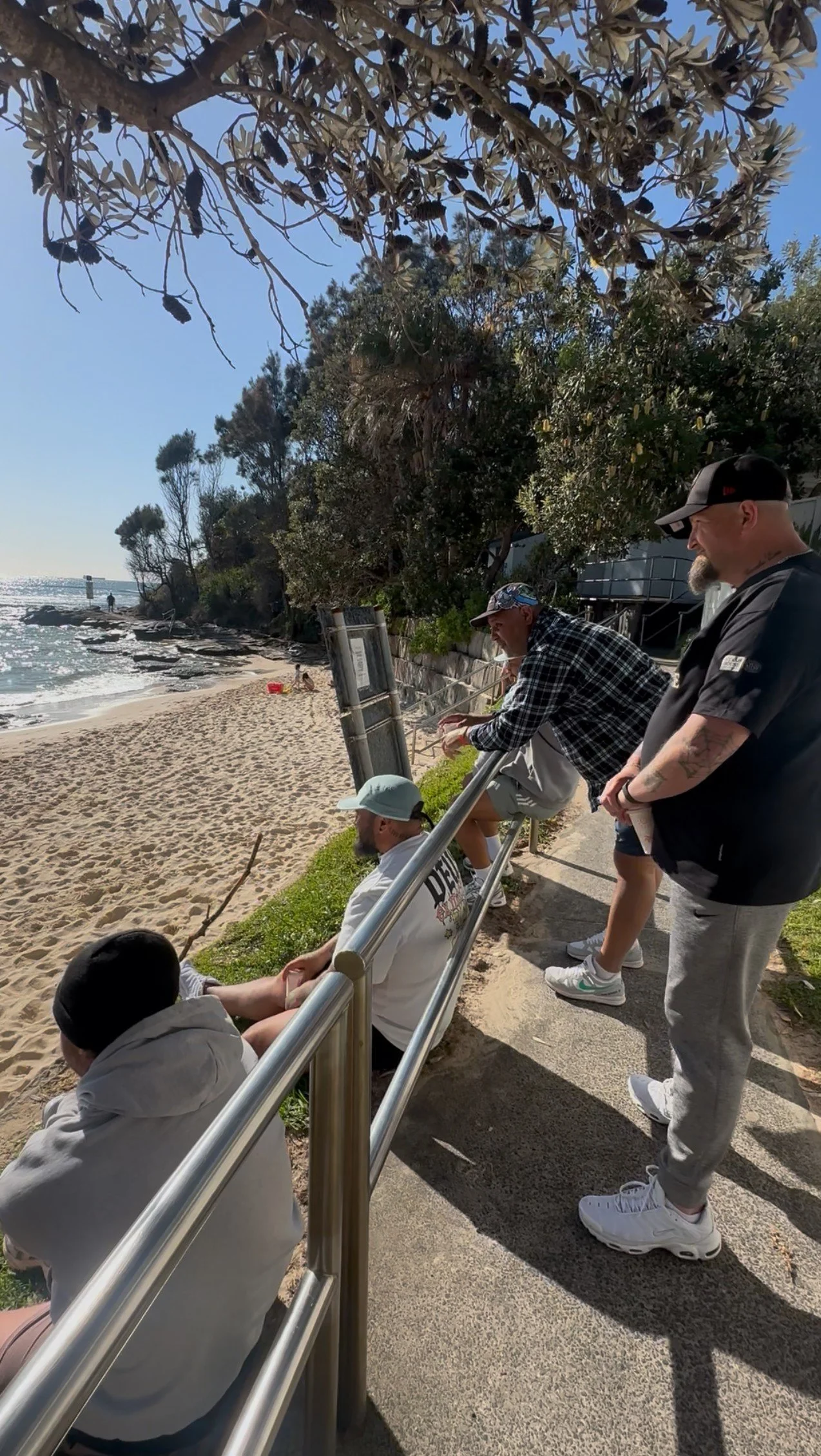 People sitting and standing along a beachside railing on a sunny day with trees in the background, near the ocean shoreline.
