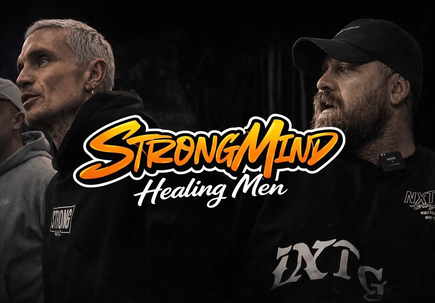 Two men sitting side by side with a black background, one has short gray hair, earrings, and tattoos, the other has a beard, mustache, and is wearing a black baseball cap. Overlaid on the image is the text 'StrongMan Healing Men' in orange and white font.