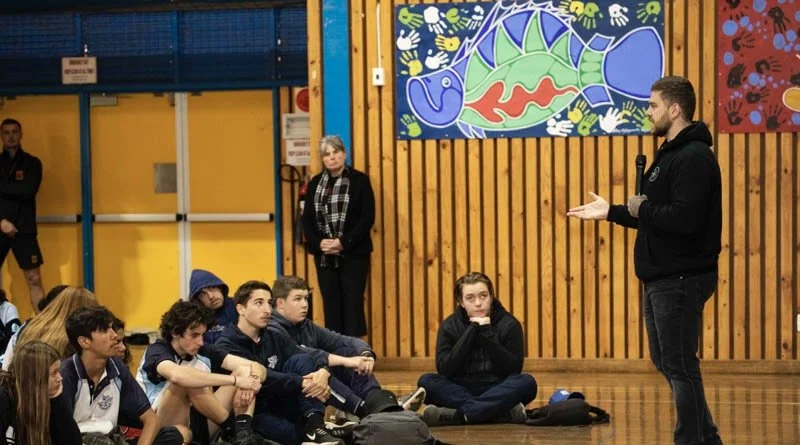 A man giving a presentation to students in a gymnasium with wooden walls and colorful artwork of a fish and other designs.