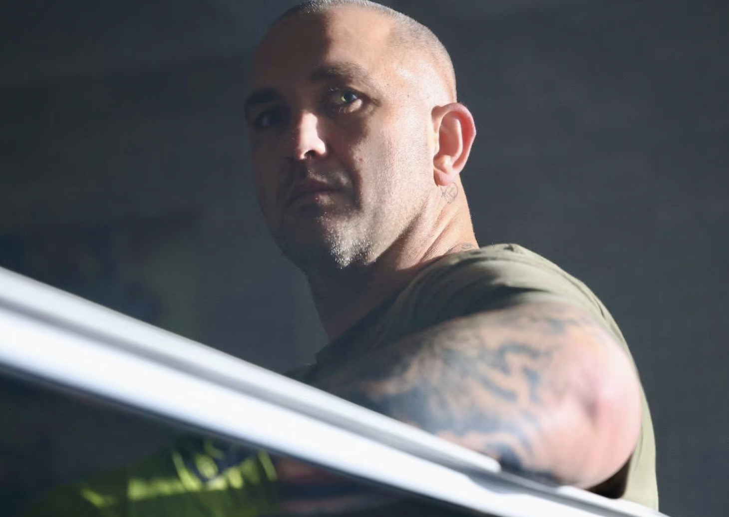 A man with a shaved head, tattoos on his arm, and a serious expression, wearing a sleeveless shirt, looking over a counter or surface against a dark background.
