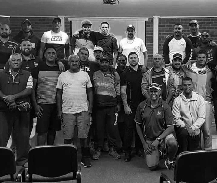 Group of men gathered indoors for a photo, standing and kneeling in front of a brick wall, with chairs at the bottom of the image.