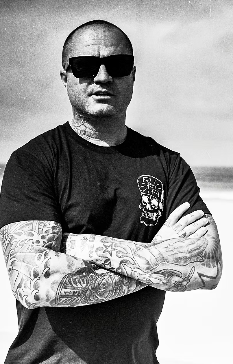 Black and white photo of a man with sunglasses, tattoos on his arms, crossing his arms, wearing a black T-shirt with a skull graphic, outdoors with an ocean or sea in the background.