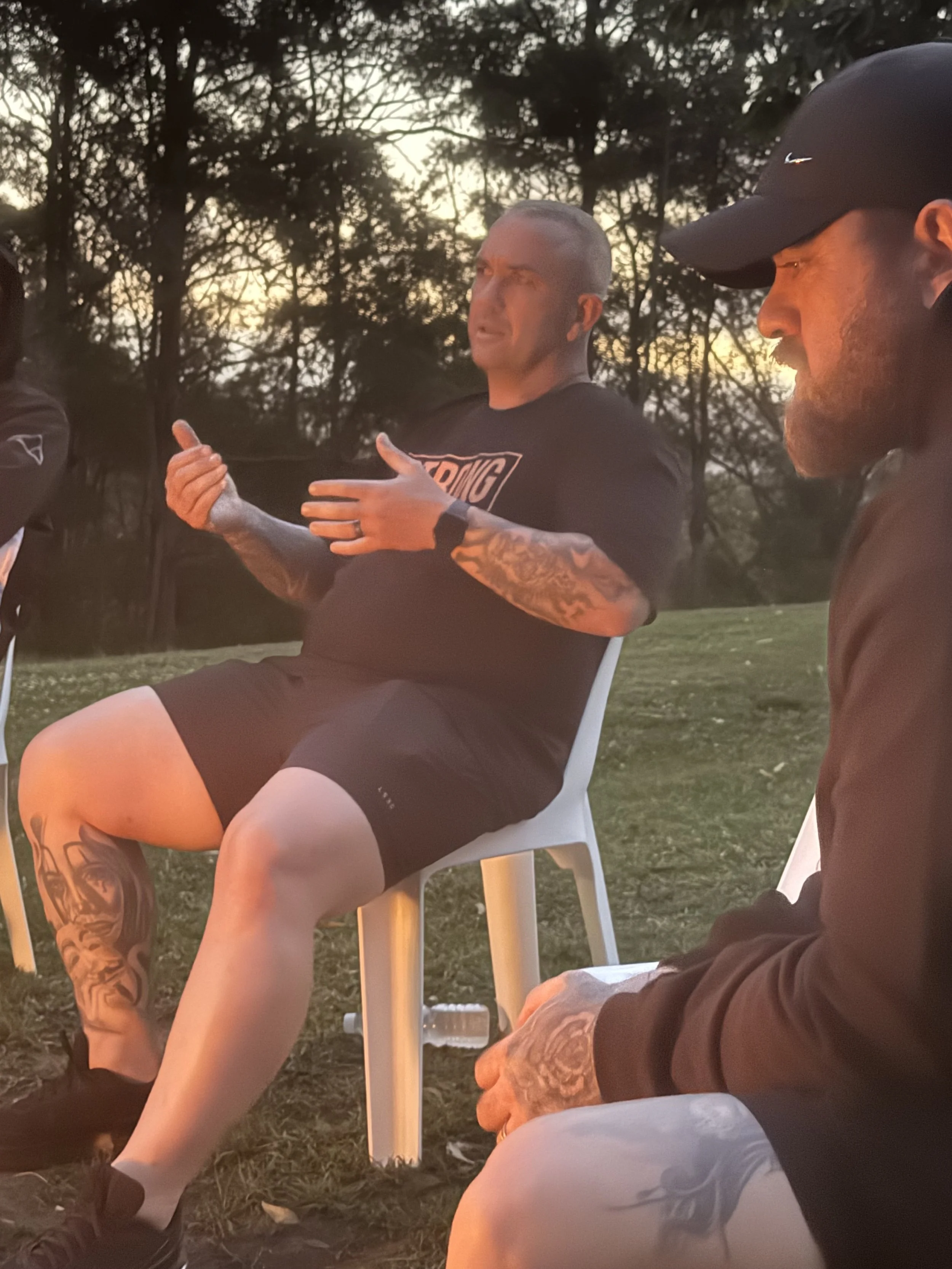 Three men sitting outdoors at sunset or early evening, with trees in the background. One man with tattoos and a shaved head is speaking or explaining, while the other man in a black cap and brown jacket listens.