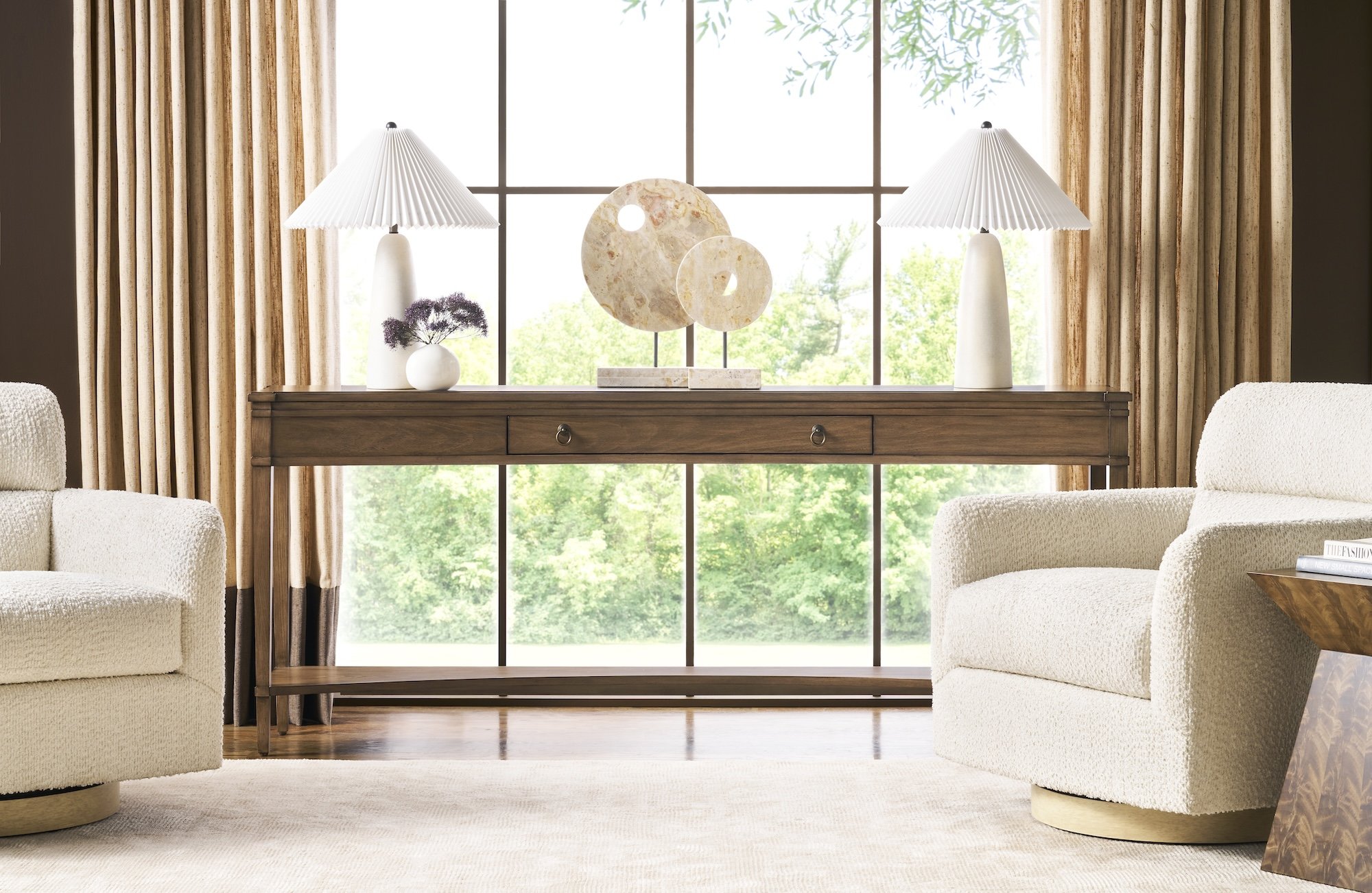 Quiet luxury living room vignette with two cream bouclé swivel chairs, a warm wood console table, sculptural stone decor, soft neutral rug, and layered drapery framed by large windows and natural light.