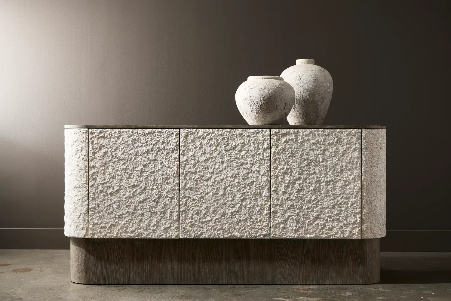 A modern textured white stone cabinet with two decorative pottery vases on top, against a dark grey wall.