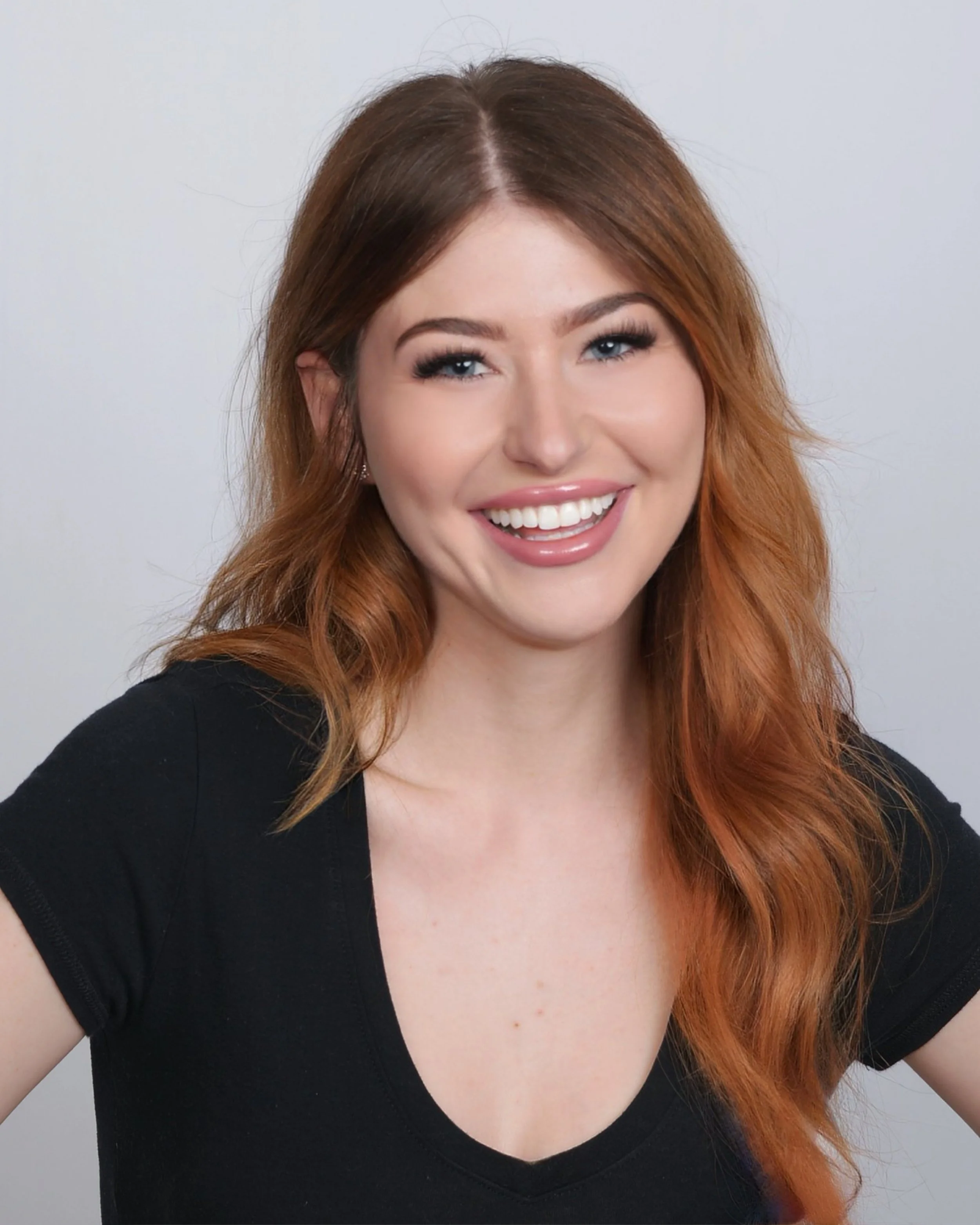 A young woman with red hair smiling at the camera, wearing a black top, against a plain gray background.