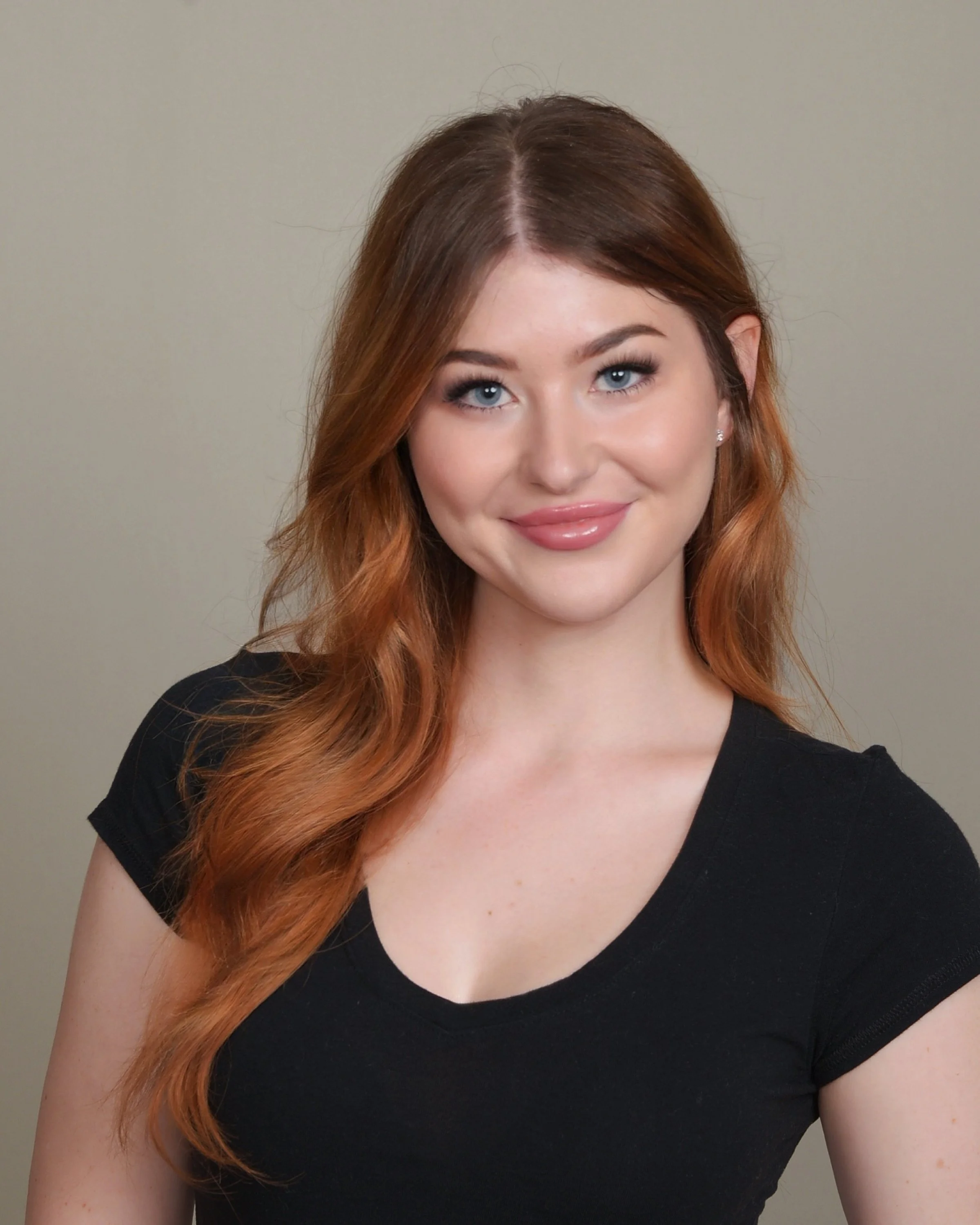 Portrait of a young woman with long wavy red hair, blue eyes, light makeup, wearing a black t-shirt, smiling against a plain background.