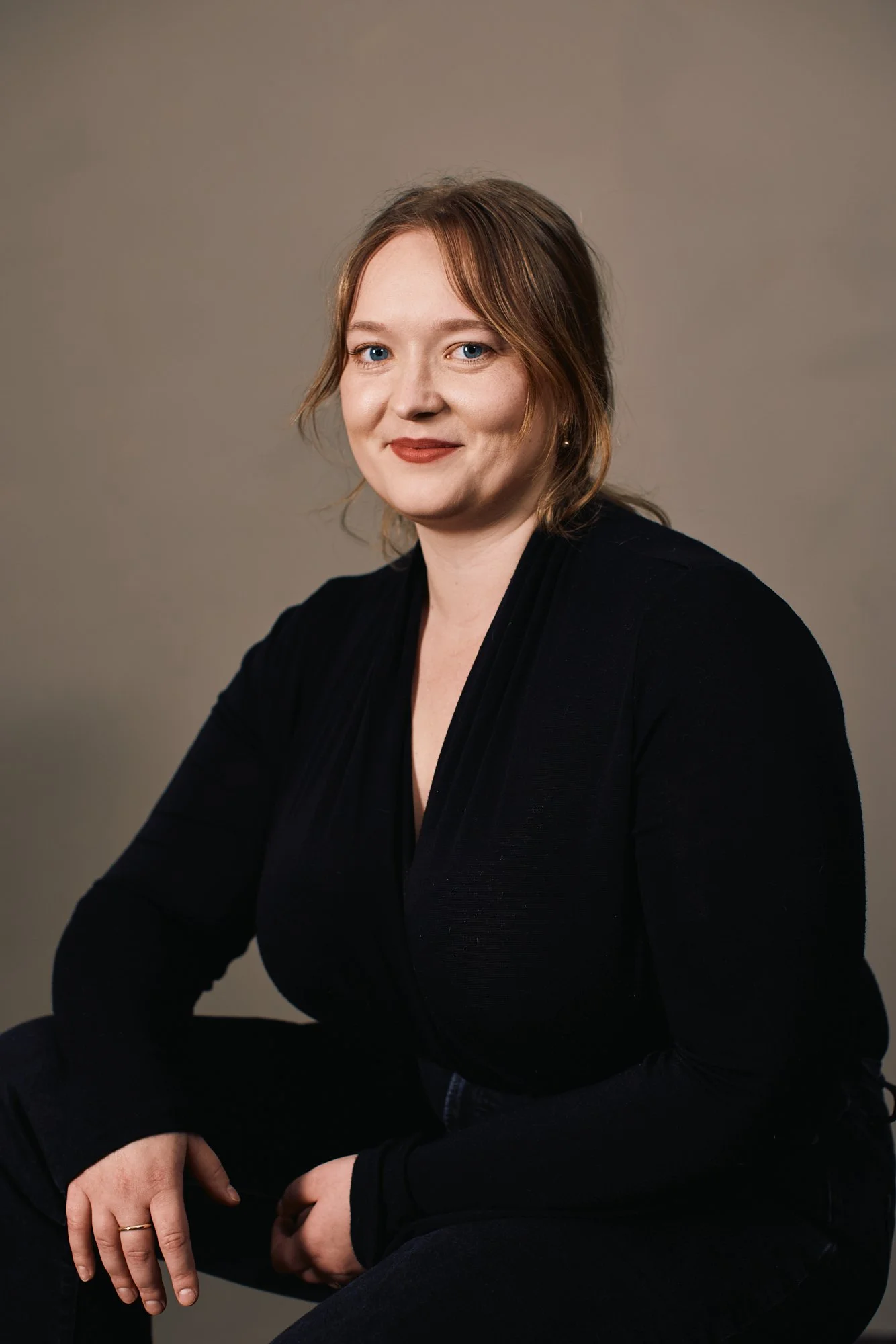 A woman with shoulder-length red hair, wearing a black top, sitting against a plain gray background, smiling at the camera.
