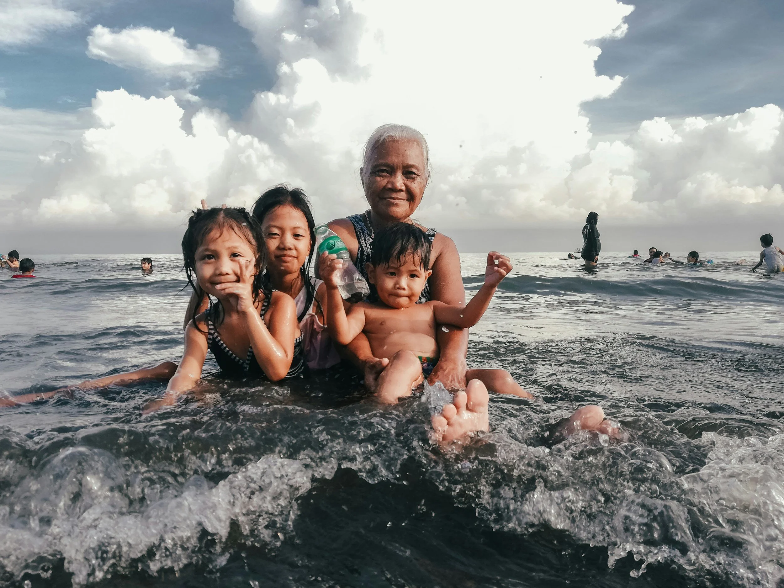 An elderly woman and three children enjoying time in the ocean on a cloudy day, with other beachgoers in the background.