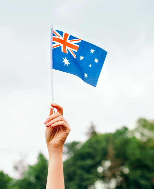 A person's hand holding an Australian flag against a background of trees and cloudy sky.