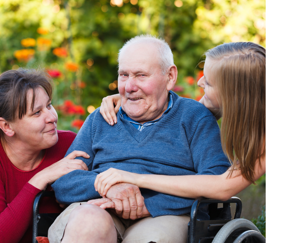 An elderly man in a wheelchair surrounded by two women, one on each side, all smiling and outdoors with colorful flowers and greenery in the background.