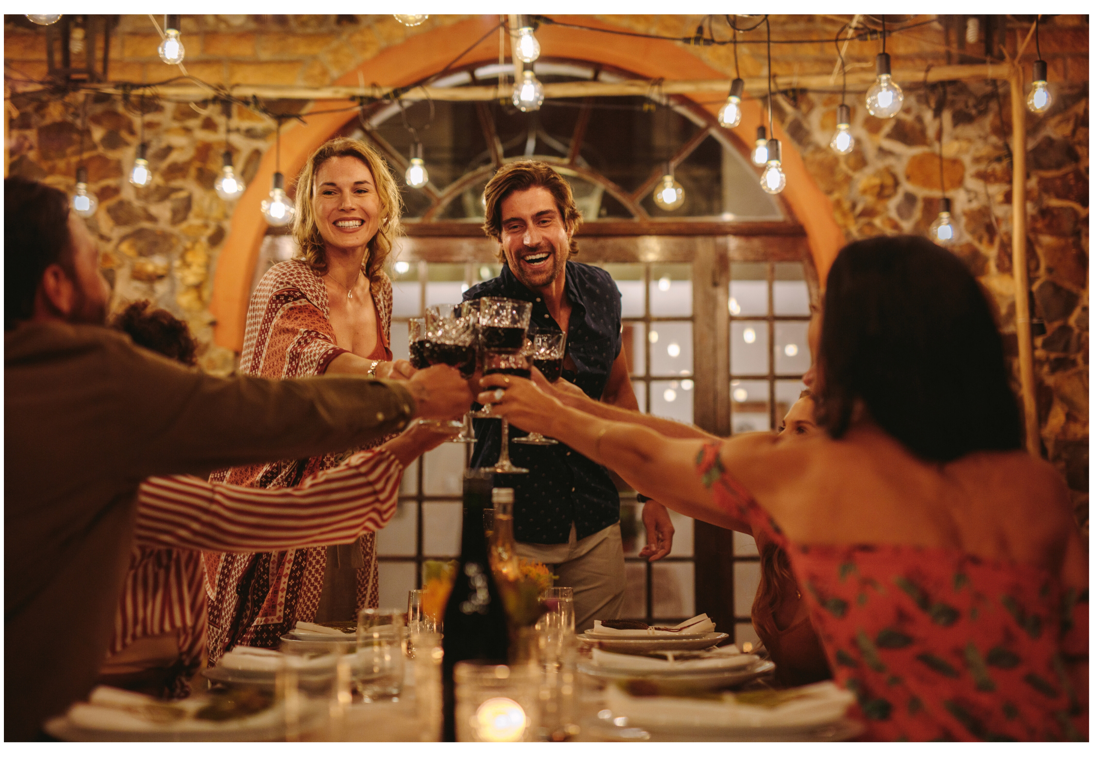 Group of friends celebrating with drinks at a dinner party, clinking glasses, in a warmly lit rustic restaurant with exposed brick walls and string lights.