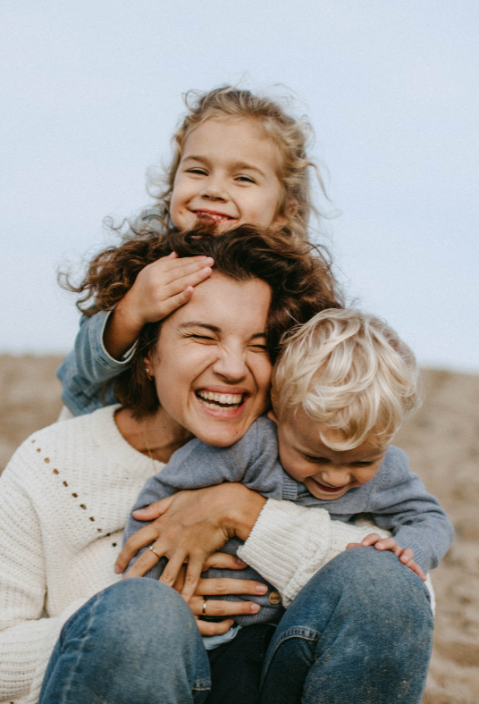 A woman with two young children, one girl and one boy, all smiling and enjoying a playful moment outdoors.