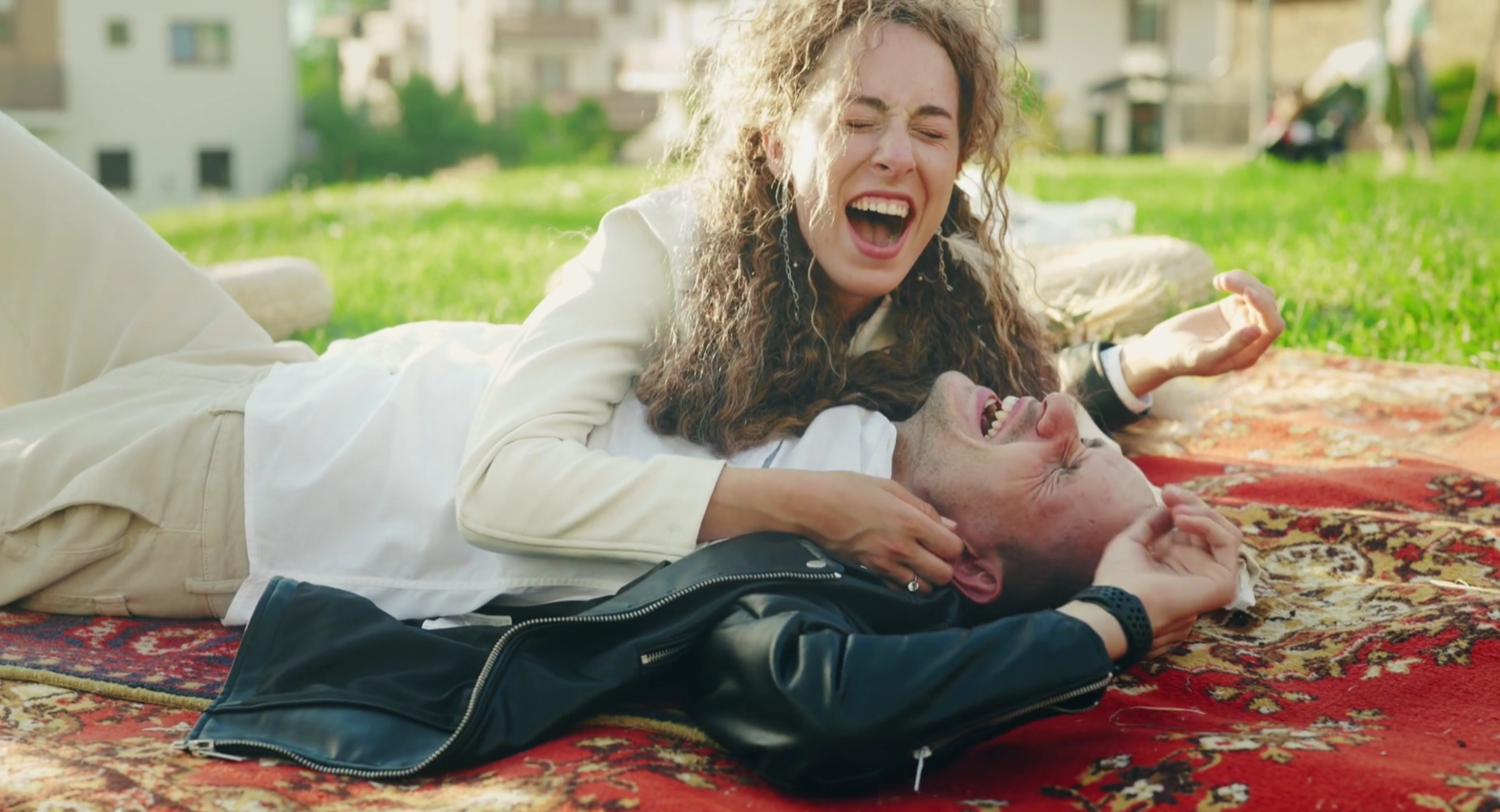 A woman with curly hair and a man with short hair and a beard are lying on a patterned rug outside in a grassy area, laughing and playfully interacting.