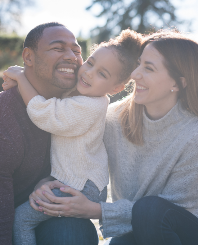 A family of three outdoors smiling and hugging, with sunlit trees in the background.