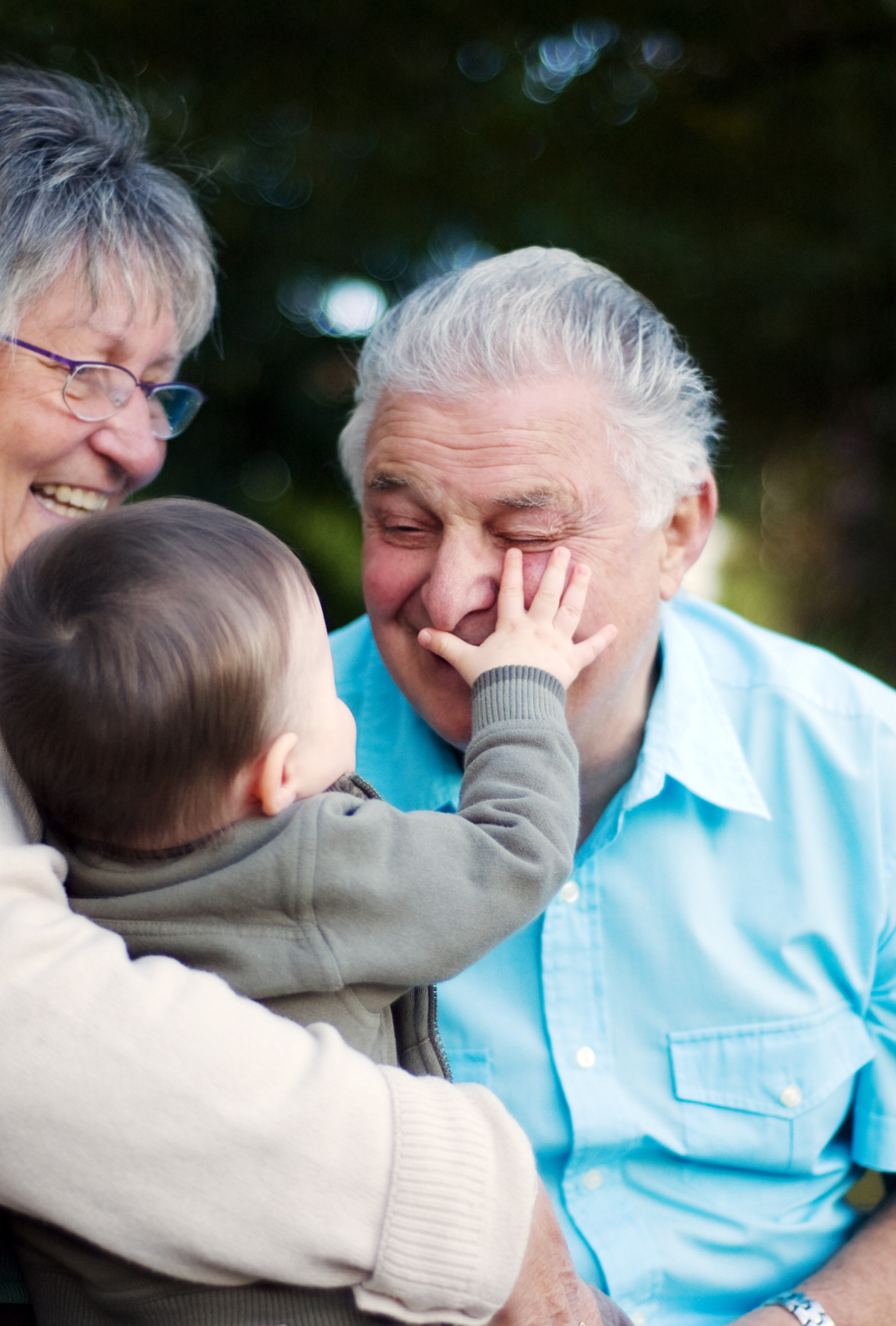 A young child touching an elderly man's face while smiling and surrounded by two women, all outdoors with greenery in the background.