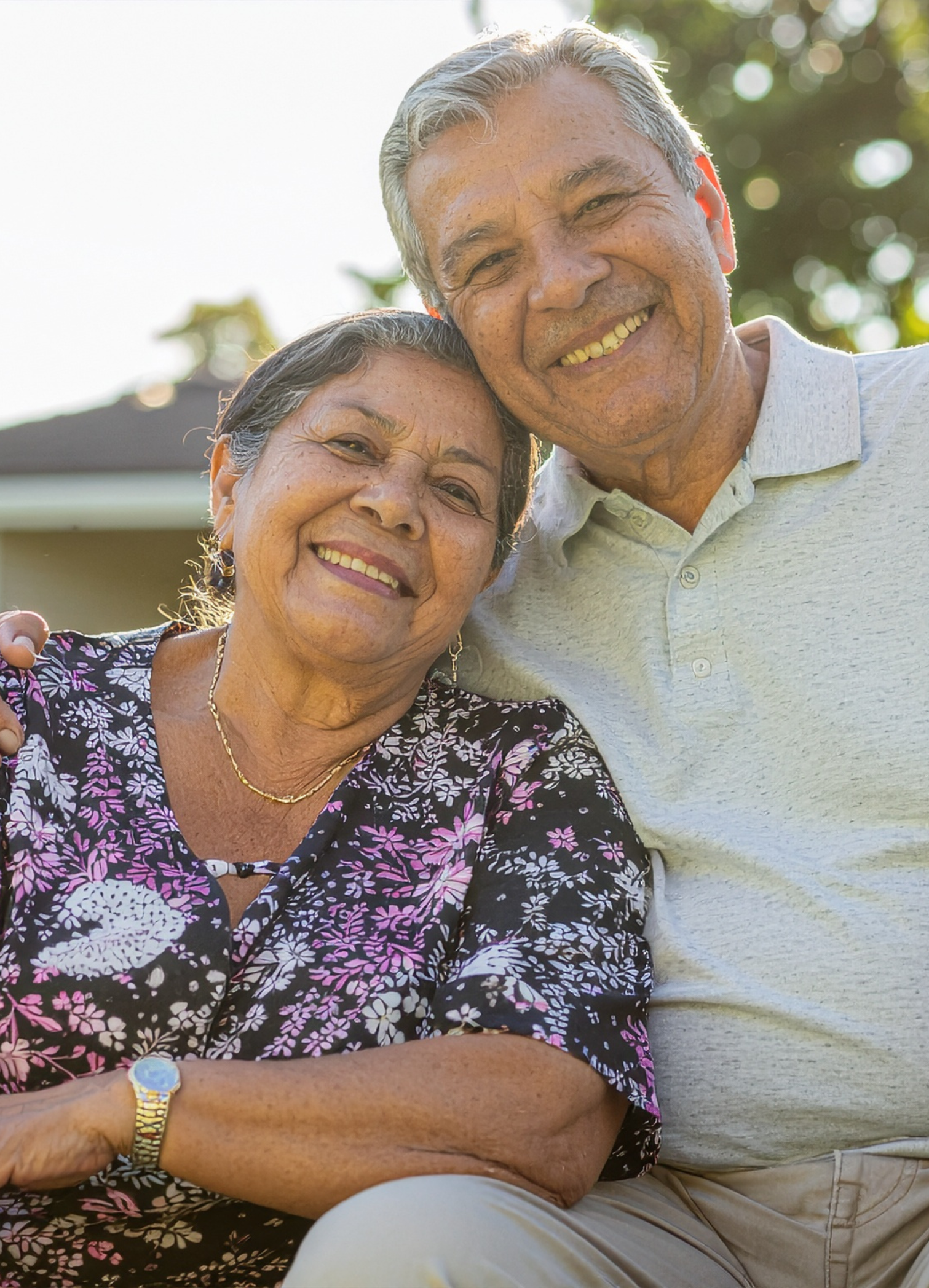 An elderly couple smiling outdoors, close-up portrait, sunny day, with a background of trees and sky.