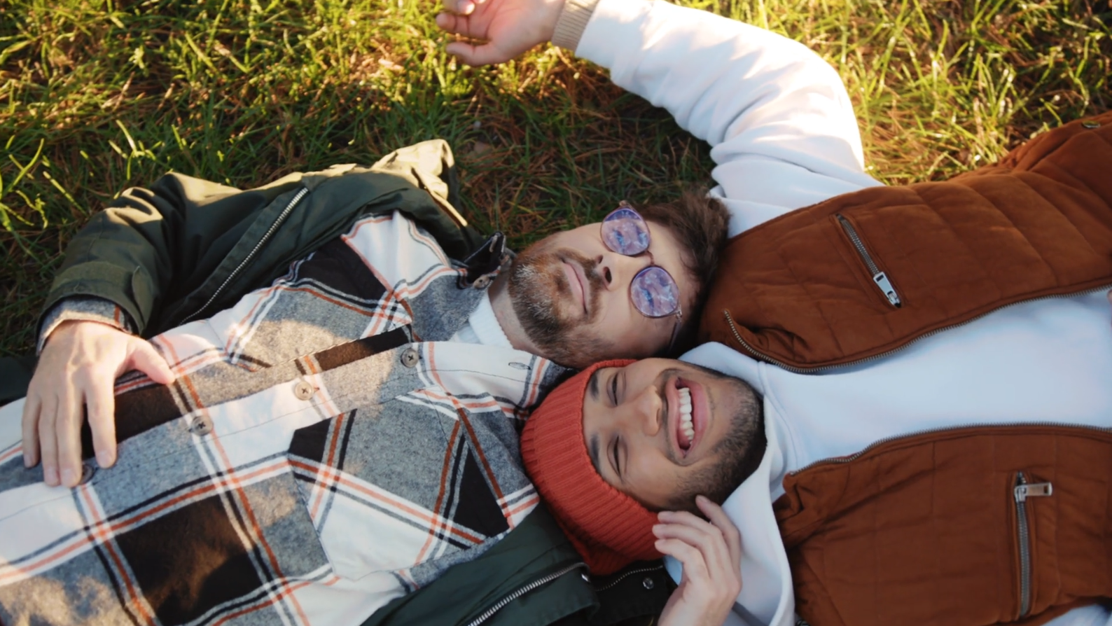 Two men lying on the grass outdoors, smiling and relaxing. One man wears glasses and a plaid shirt with a dark jacket, while the other wears a red beanie and a brown vest. They appear happy and content.