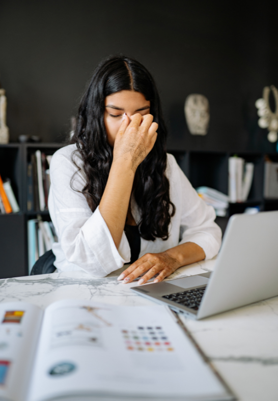 A woman with long dark hair sitting at a desk, covering her face with her hand, in front of a laptop and an open book, in a modern home or office space.