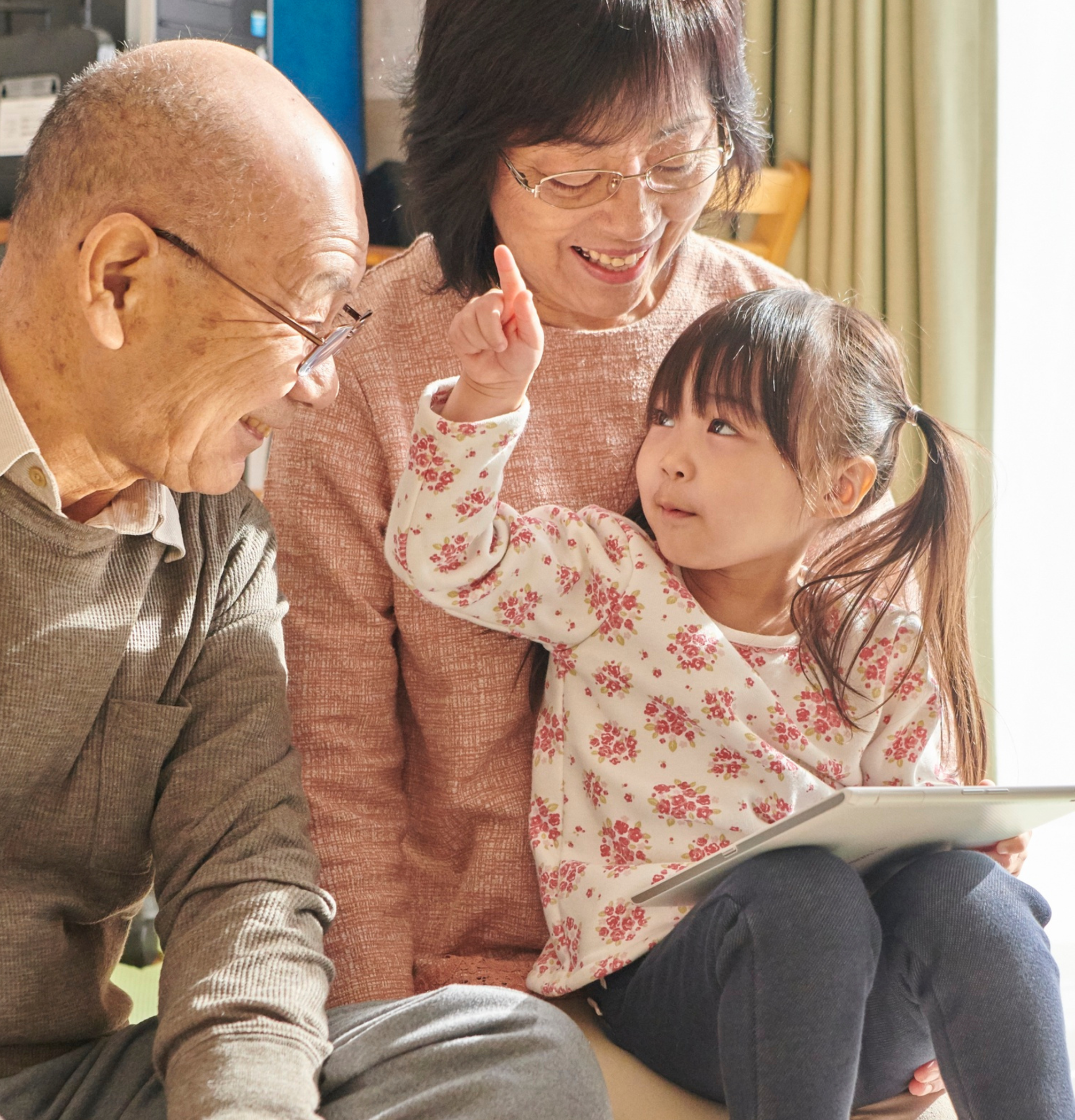 Three generations of family members smiling and interacting indoors, with a young girl showing a hand gesture while sitting with an older man and a middle-aged woman, all engaging happily.