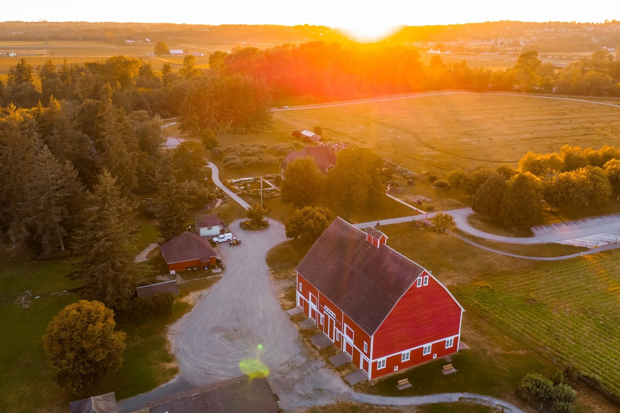 Aerial view of a farm at sunset featuring a large red barn, smaller buildings, winding gravel roads, green fields, and surrounding trees with sunlight casting a warm glow.