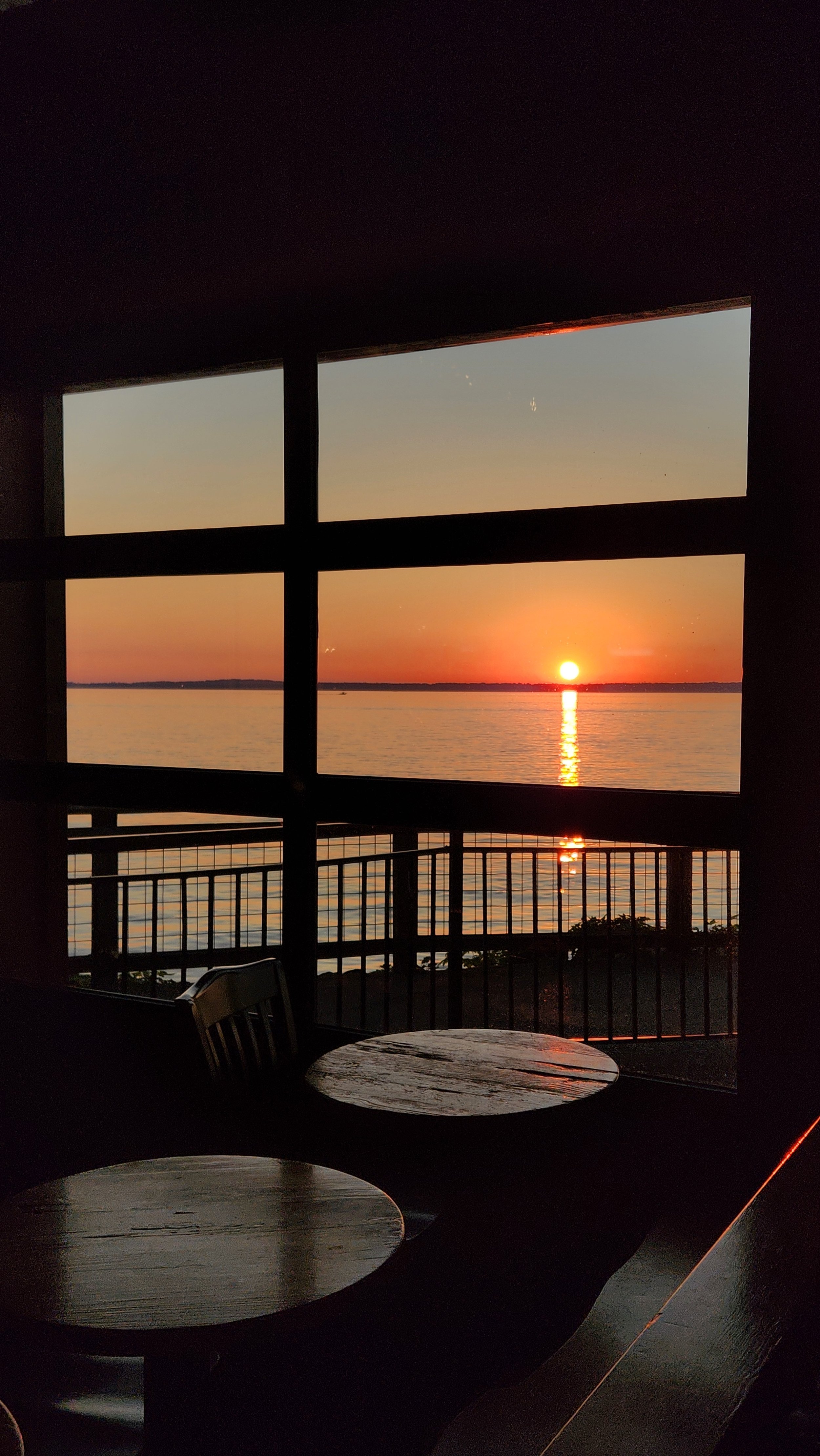 Sunset over a calm body of water viewed through large window panes, with a wooden table and chairs inside.