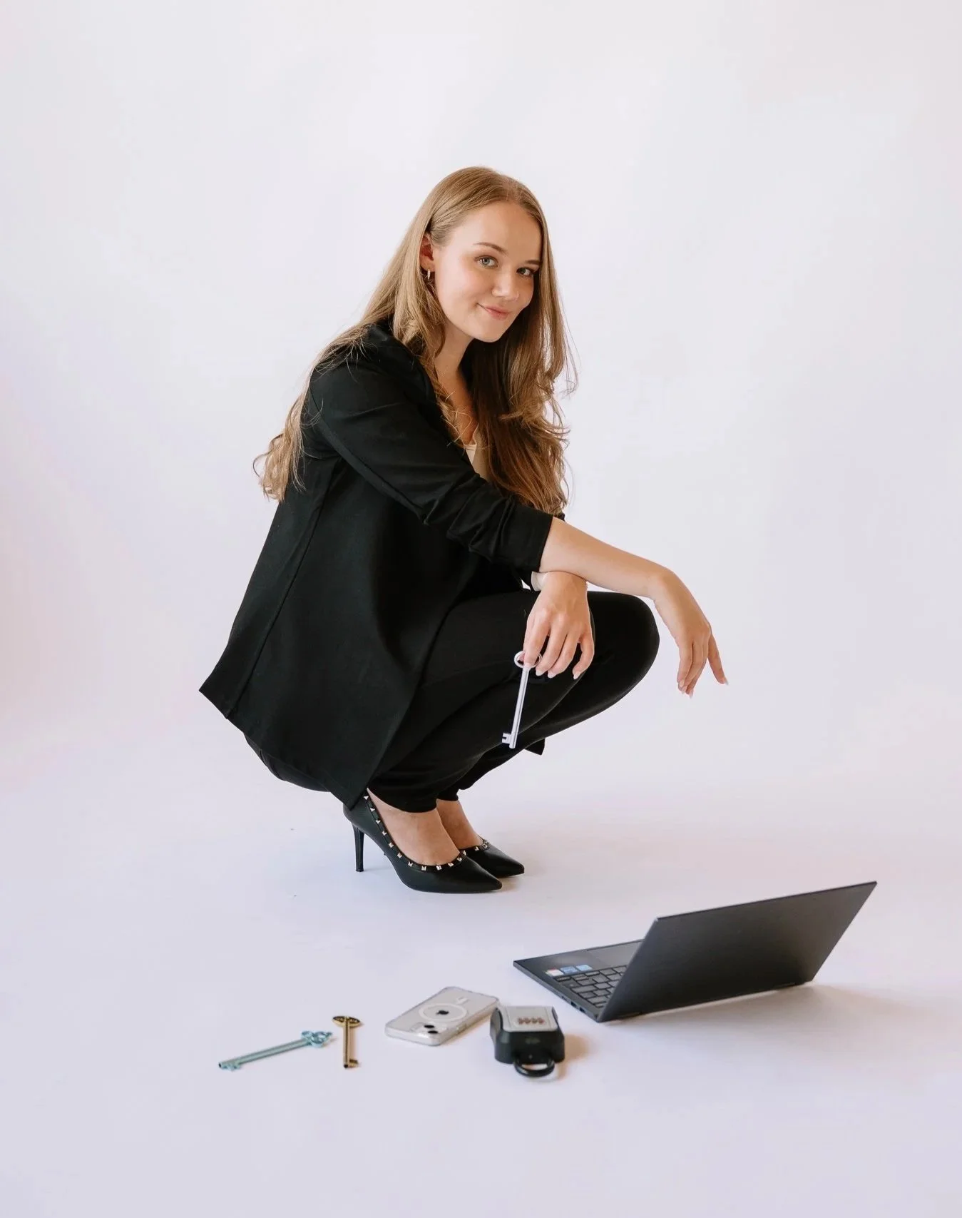 A woman in black high heels and a black blazer squats with her legs spread in front of a laptop, looking at the camera and smiling. Items on the floor include two keys, a phone, a camera, and an open laptop against a plain white background.