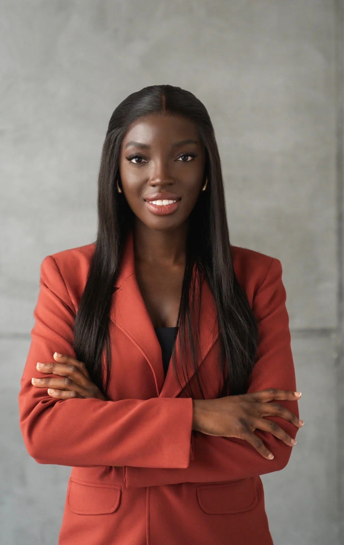 A professional African American woman with long black hair, wearing a rust-colored blazer and a black top, standing with arms crossed against a gray background.