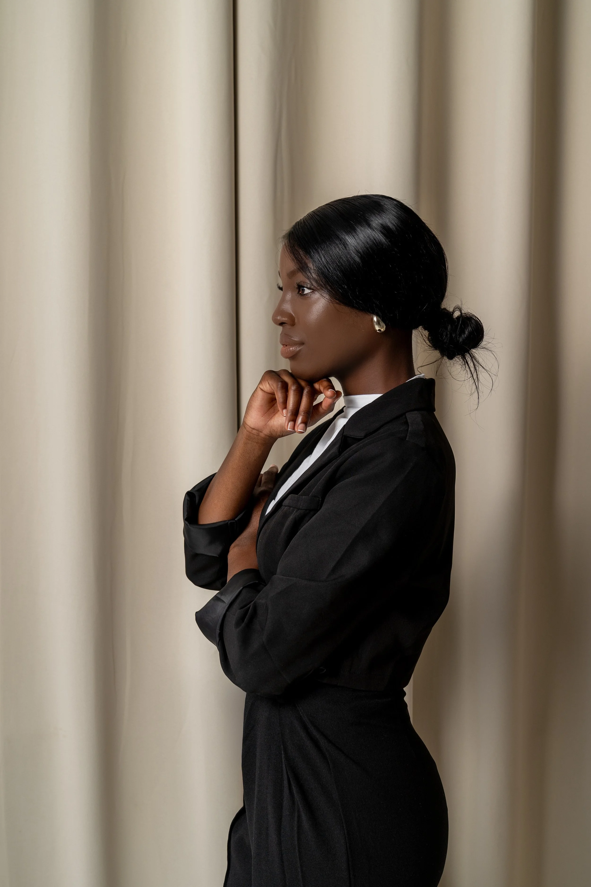 A woman with black hair styled in a low bun, wearing a black blazer and earrings, standing in profile against a cream-colored curtain.