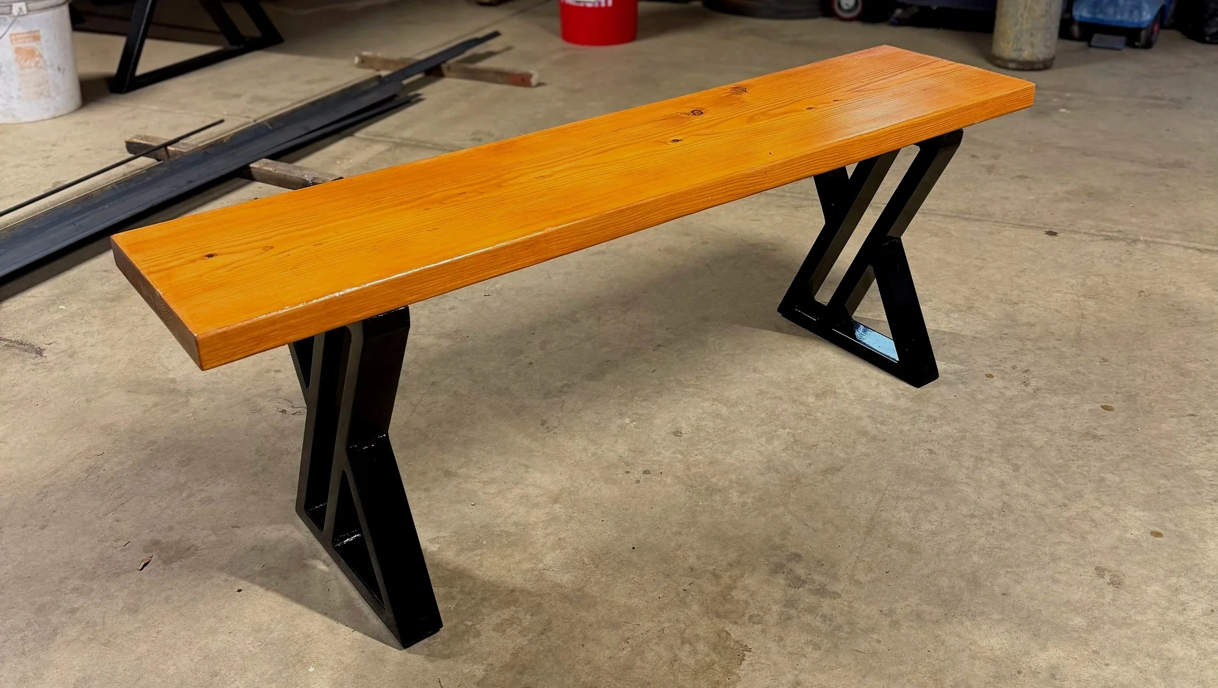 Wooden bench with black metal X-shaped legs placed on concrete floor in a workshop or garage setting.