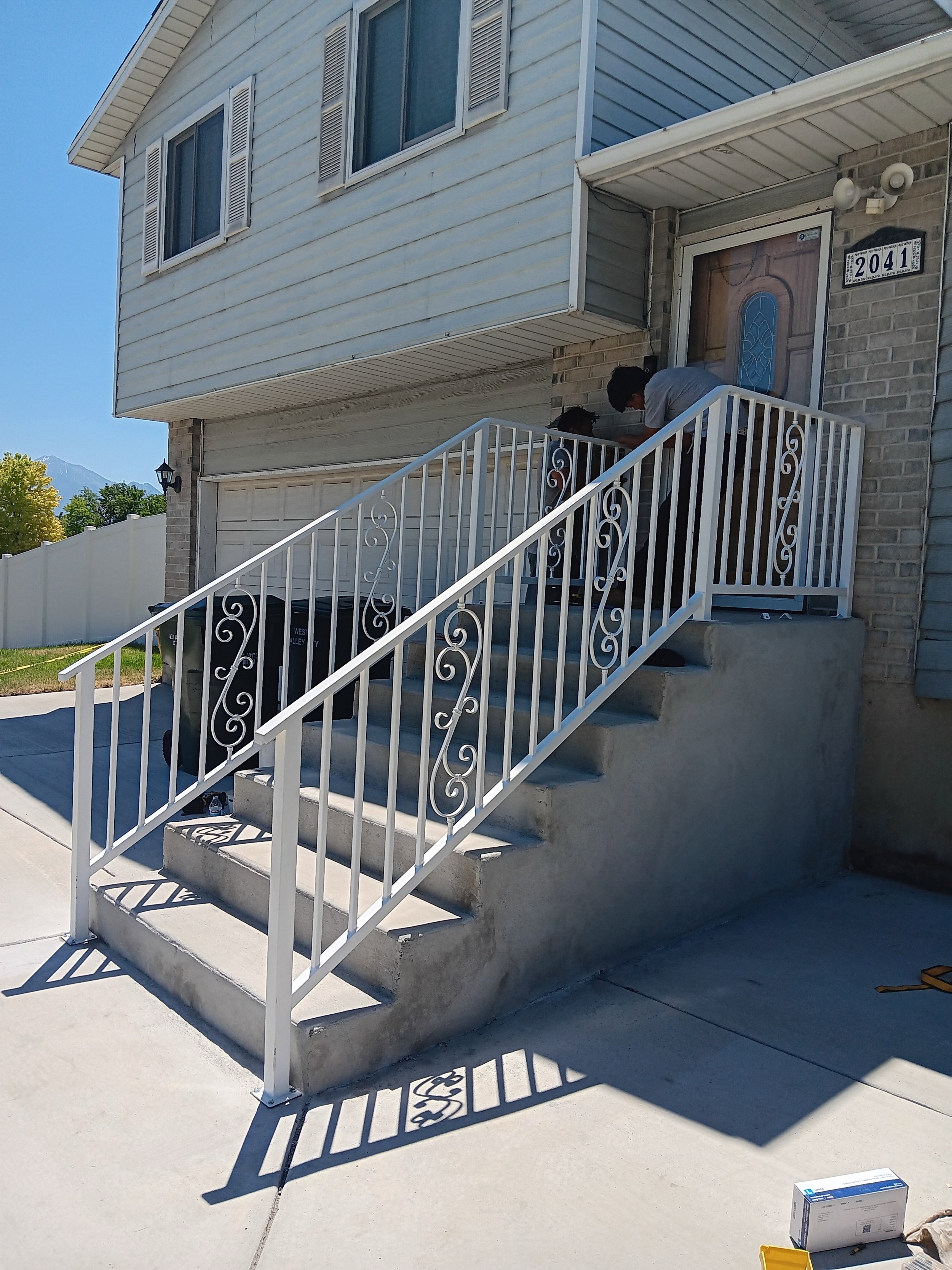 People working on a front porch of a house with stairs and white railing, with a trash can near the house