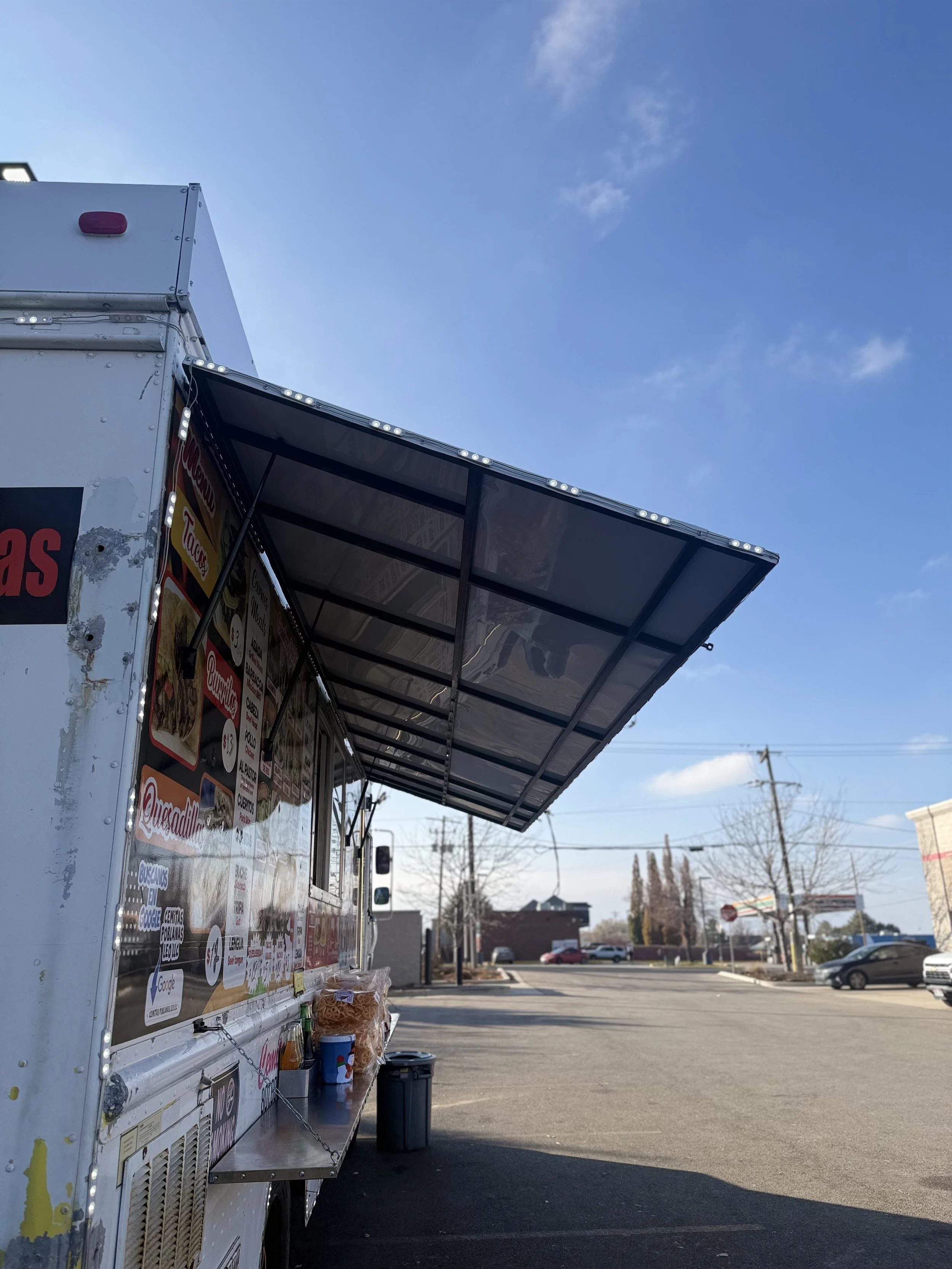 A food truck parked in a lot with clear blue skies and some clouds, advertising menus on its side, and some food items visible.