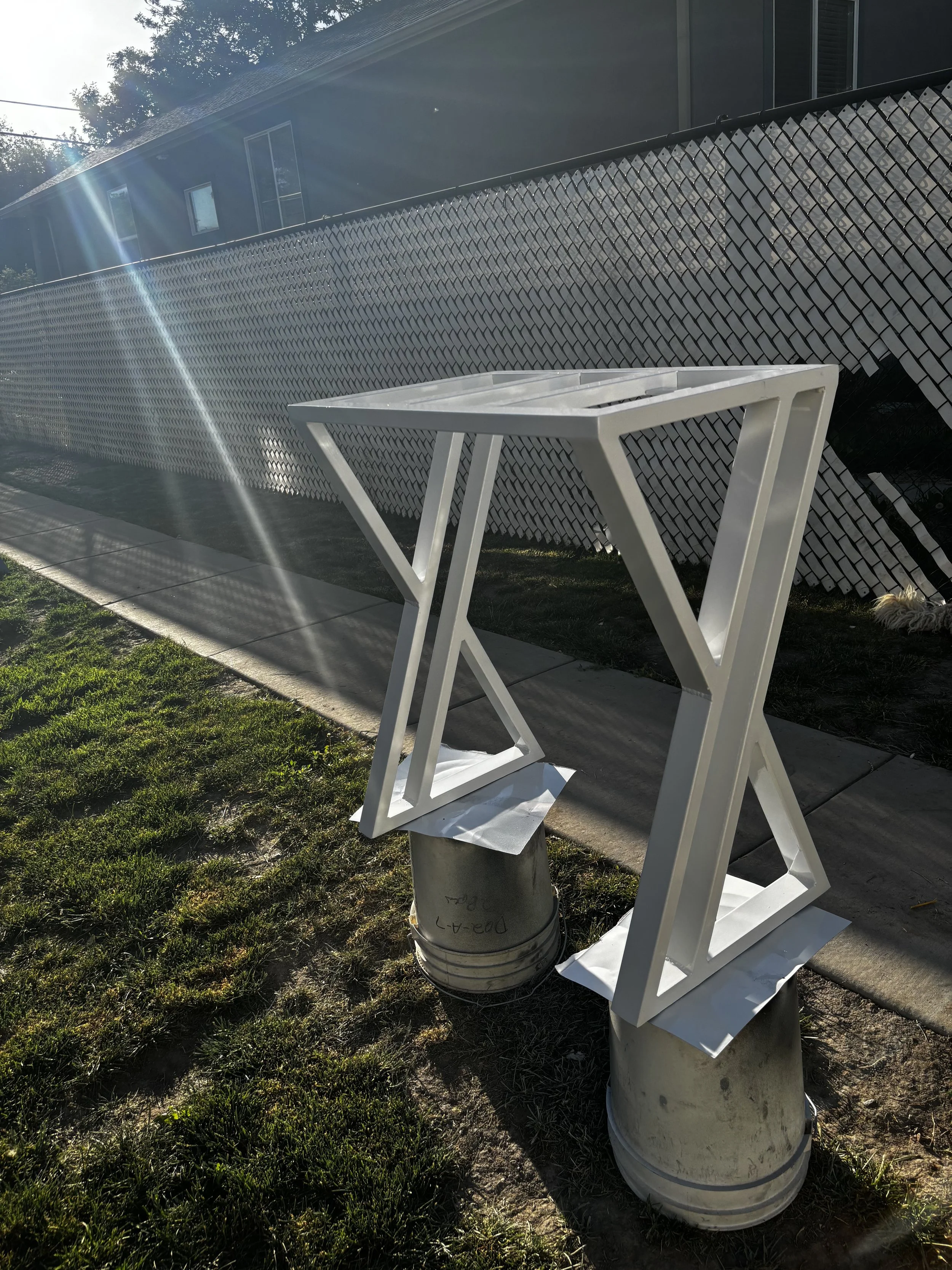 A white plastic table placed on top of metal buckets in a backyard with green grass, with a house, window, and a white lattice fence in the background. Sunlight creates lens flare in the image.